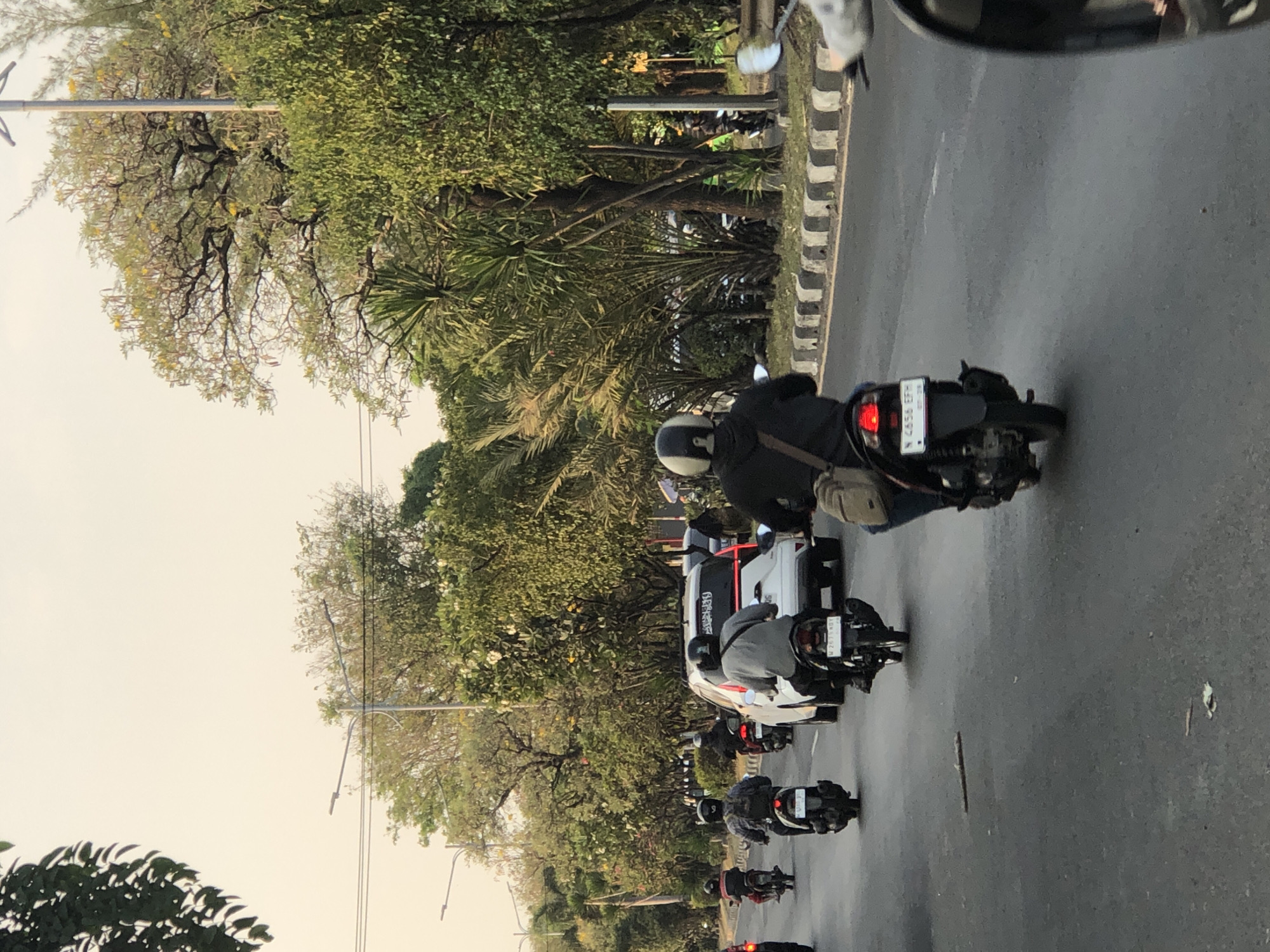 The image depicts a street scene in Ngaban, Indonesia, under a bright, hazy sky. Several motorcycles are in motion, with riders wearing helmets. A white car is also present on the road. The environment is lined with trees and vegetation. The road has distinct markings and a curb with alternating black and white stripes. The overall impression is of a typical day in a city with moderate traffic.