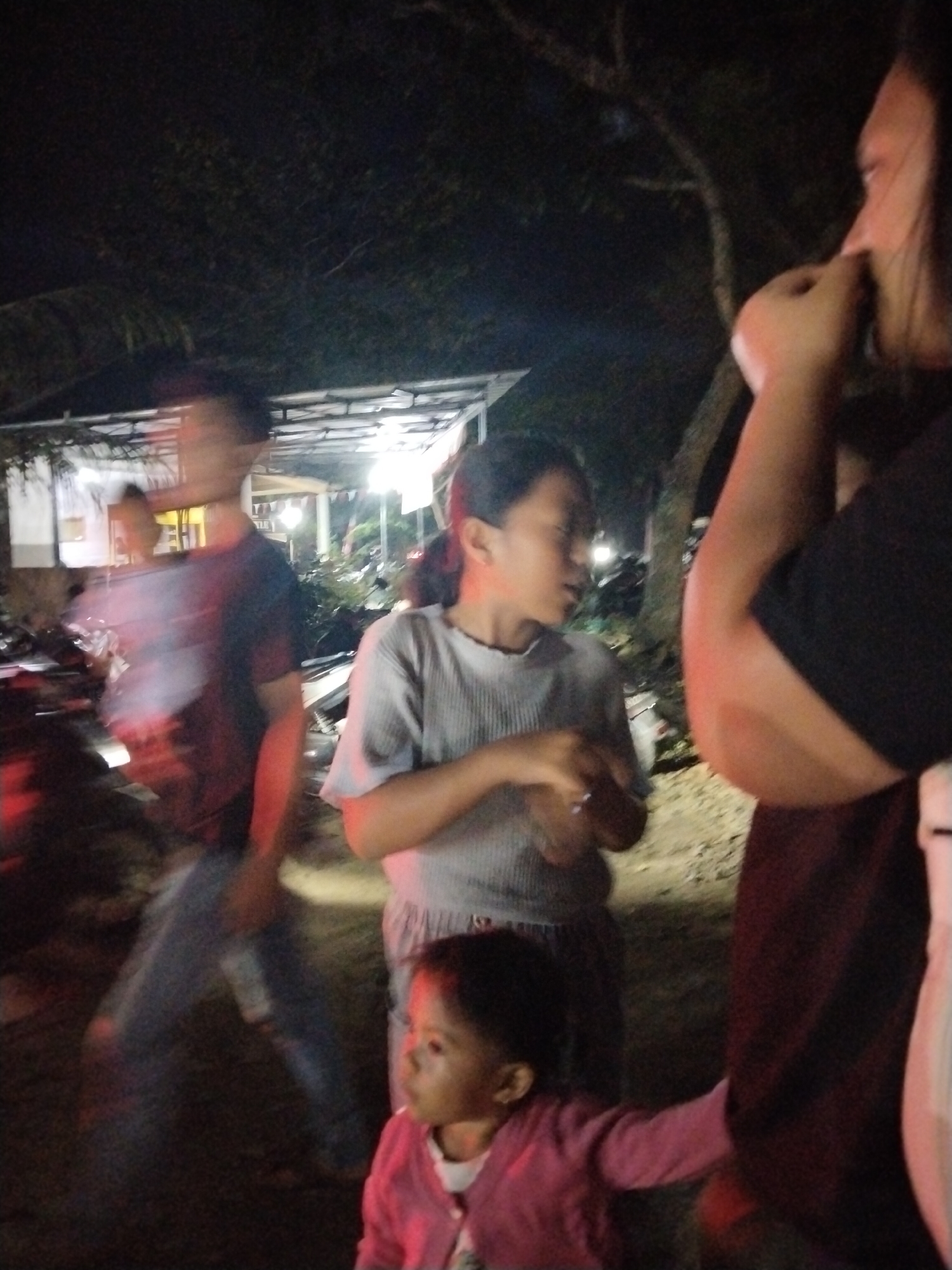 The image depicts a nighttime scene at a street-side gathering in Tasikmadu, Indonesia. The lighting is dim and appears to be artificial, casting a reddish and yellow hue on the subjects. In the foreground, a young girl wearing a pink jacket is looking towards the right of the frame. To her left, a person wearing a light grey, ribbed shirt is also present. Behind them, a blurred figure in a dark t-shirt is visible, and further back, another blurred individual. The background consists of dimly lit trees, foliage, and what appears to be a building structure with a corrugated roof, possibly a stall or shop. The ground is covered in dirt and small stones. The overall impression is of a casual, outdoor community event.