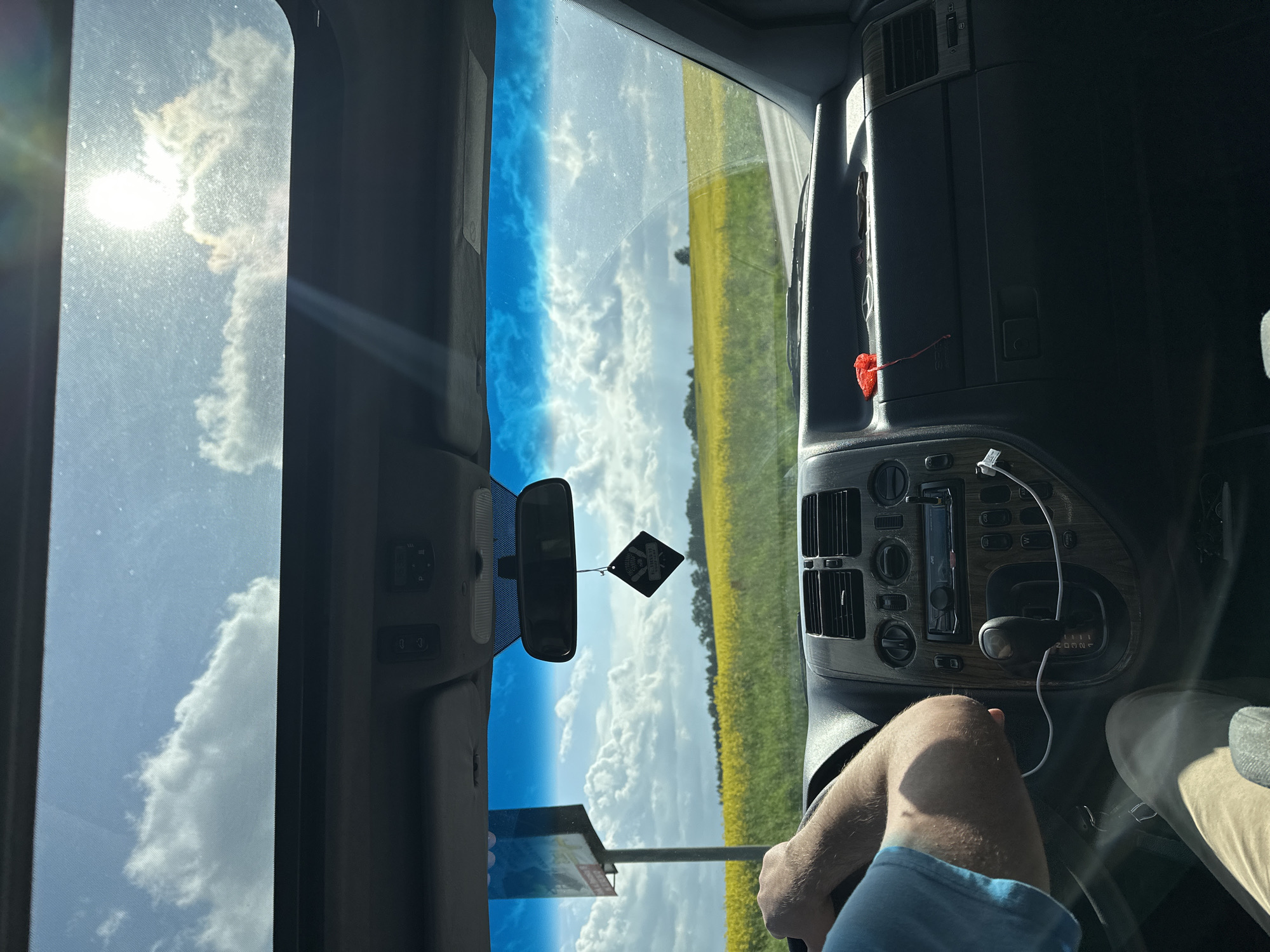 A view from inside a vehicle, potentially a car or van, moving on a road. The landscape visible through the windshield and sunroof includes a field of yellow flowers, likely rapeseed or a similar crop, alongside a stretch of road. The sky is blue with scattered clouds. The interior of the vehicle shows the dashboard, steering wheel, gear shift, and part of the driver's arm and leg. There is an air freshener hanging from the rearview mirror and a red object dangling nearby the center console. The location is Rzepowskie, Poland, indicated by a road sign visible through the windshield.