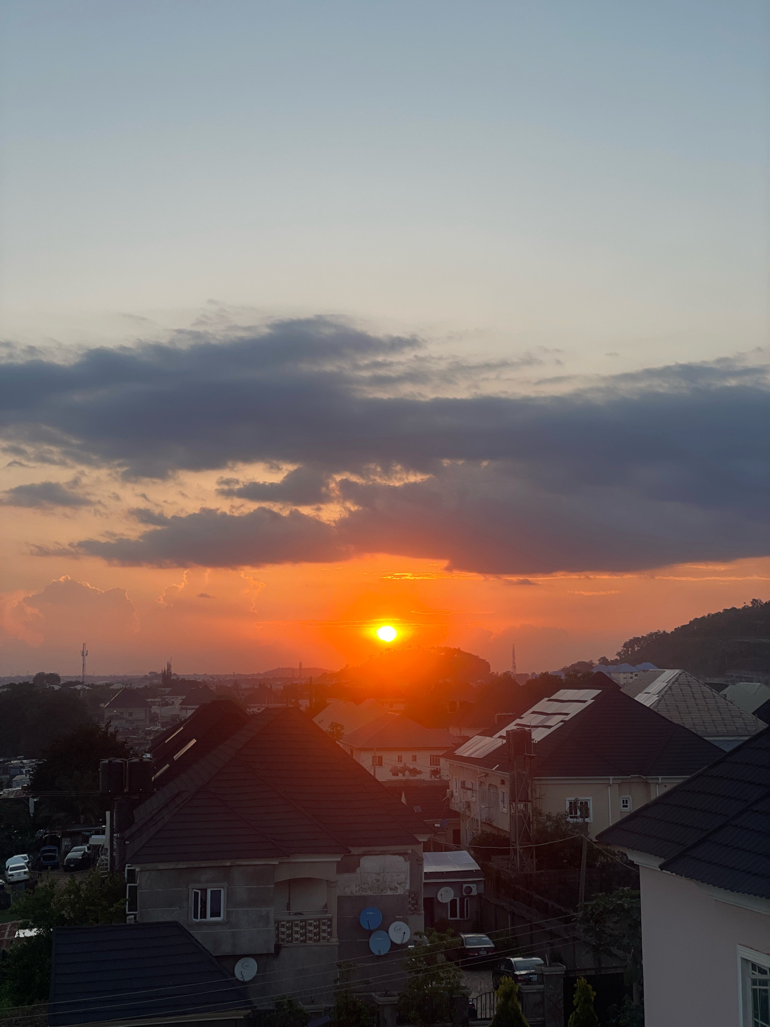 This is a photograph of a sunset over a residential area in Abuja, Nigeria. The sun is a bright orange orb low on the horizon, casting a warm glow on the scattered clouds. The sky transitions from a pale blue at the top to a vibrant orange near the sun. In the foreground, several houses with dark, pitched roofs are visible. Some of these houses have visible solar panels and water tanks. A hill covered in trees and vegetation is visible in the background to the right, with more houses scattered on its slope. To the left of the frame, a few vehicles are parked, and a telecommunications tower stands in the distance. The overall scene depicts a typical evening in a suburban or semi-urban environment in Abuja.