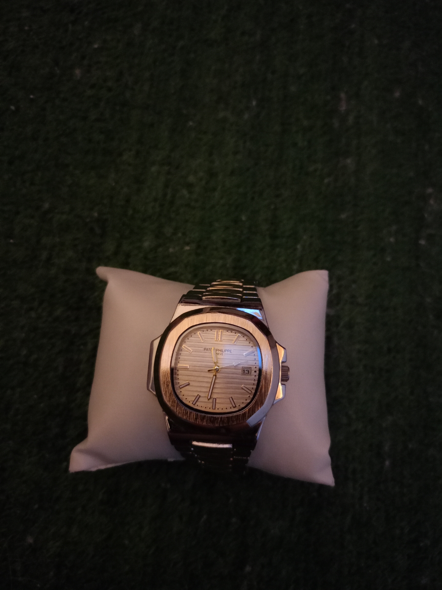 A wristwatch with a metal band and a champagne-colored face with horizontal lines and gold hour markers is displayed on a white, square pillow. The watch has the brand name "PATEK PHILIPPE GENEVE" printed on the face, and a date window showing "13". The pillow is placed on a dark green, textured surface that resembles artificial turf. The background is dark and out of focus. There are no people present in the image, nor are there any indications of the specific location within the city of Petrozavodsk, Russia.