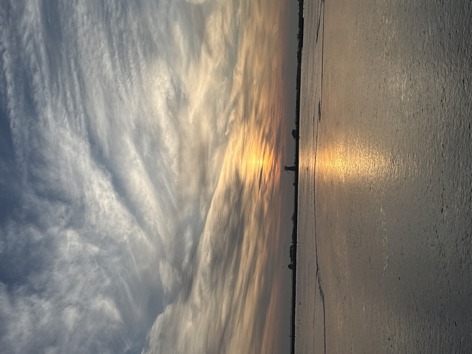 The image displays a sunset over Breydon Water, west of Great Yarmouth, United Kingdom. The sky occupies the upper two-thirds of the frame, showing white, wispy cirrus clouds against a pale blue background, gradually transitioning to orange and yellow hues closer to the horizon. The setting sun, a bright orange-yellow sphere, is positioned low on the horizon, slightly to the right of the central vertical axis. Its intense light creates a prominent, reflective path across the calm water surface in the foreground, extending towards the viewer. On the distant horizon, the dark silhouette of Berney Arms Windmill with its sails is prominently visible to the right of the sun. Several smaller, dark structures are also silhouetted on the horizon to the left of the windmill. The water surface in the foreground and midground exhibits minor ripples. No human figures or animals are explicitly discernible within the scene.