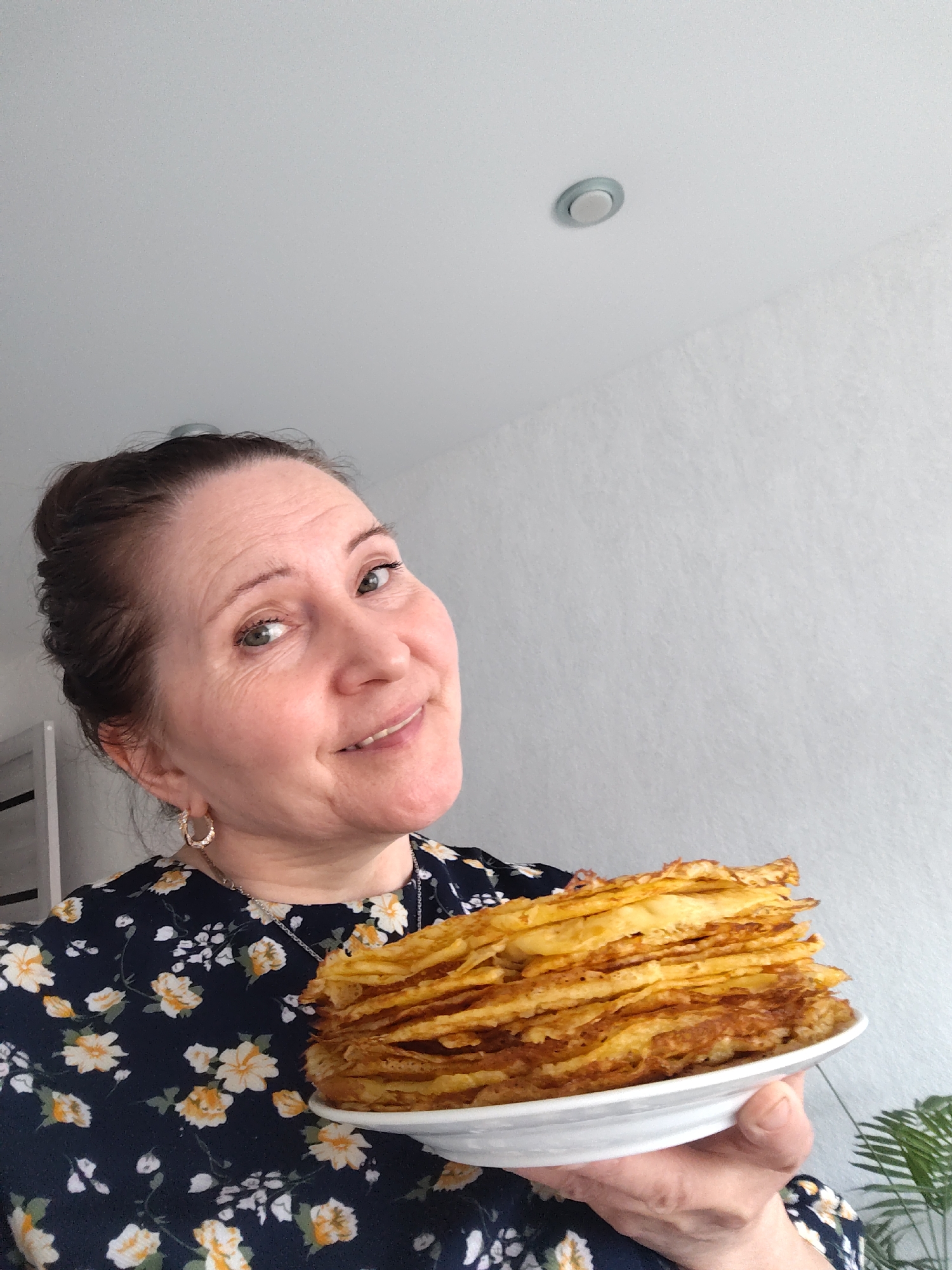 A woman is captured in a close-up, indoor shot, holding a substantial stack of pancakes or blini on a white plate. She is positioned slightly to the left, looking directly at the camera with a warm, open-mouthed smile, her green eyes sparkling. Her dark hair is pulled back, revealing her forehead and face. She appears to be middle-aged, with a pleasant and content expression. She is wearing a dark blue top or dress adorned with a white and yellow floral pattern. Gold-colored hoop earrings and a delicate silver necklace are visible.
The stack of pancakes is golden-brown and appears freshly made, suggesting a large batch was prepared. The background features a plain light gray wall with a subtle textured pattern, and a portion of a white ceiling with a small, circular recessed light fixture. A hint of green foliage from a houseplant is visible on the far right, while a blurred white structure can be seen on the left. The overall impression is one of domesticity and culinary achievement, possibly celebrating a traditional meal or holiday in Sarapul, Russia. The bright, even lighting suggests it is daytime. No visible text is present in the image.