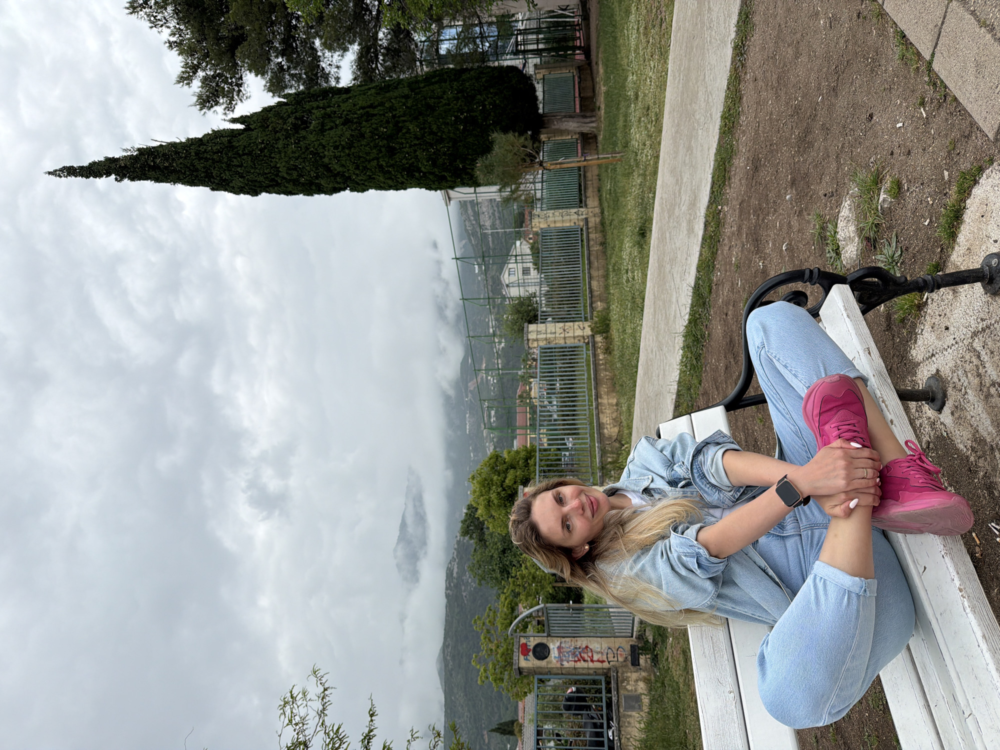 A full body shot shows a blonde woman wearing light blue jeans, a light blue jean jacket, and pink sneakers, sitting on a white bench. She is looking at the camera and has her legs crossed. In the background are trees and a mountain range obscured by fog. In the middle ground is a park with a walkway, grass, and a tall, pointed tree. Below the park is a fence, a building, and more trees. The image was taken on a cloudy day. The location is in the city of Budva, Montenegro.
