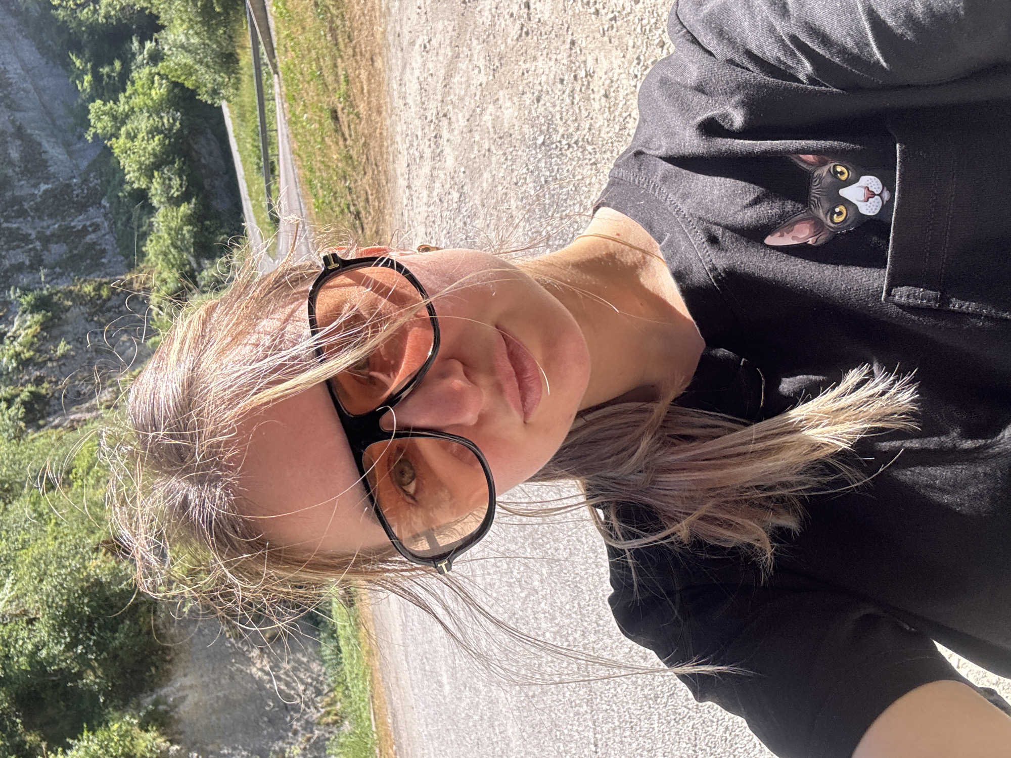 A young woman with blonde hair and sunglasses is taking a selfie outdoors. She is wearing a black t-shirt with a printed image of a cat peeking out of a pocket. The background shows a steep, rocky cliff face covered in greenery, with a winding road visible in the distance. The lighting suggests it is a sunny day. The image does not contain any information that allows for identifying the location in the city of L'Essard, France.