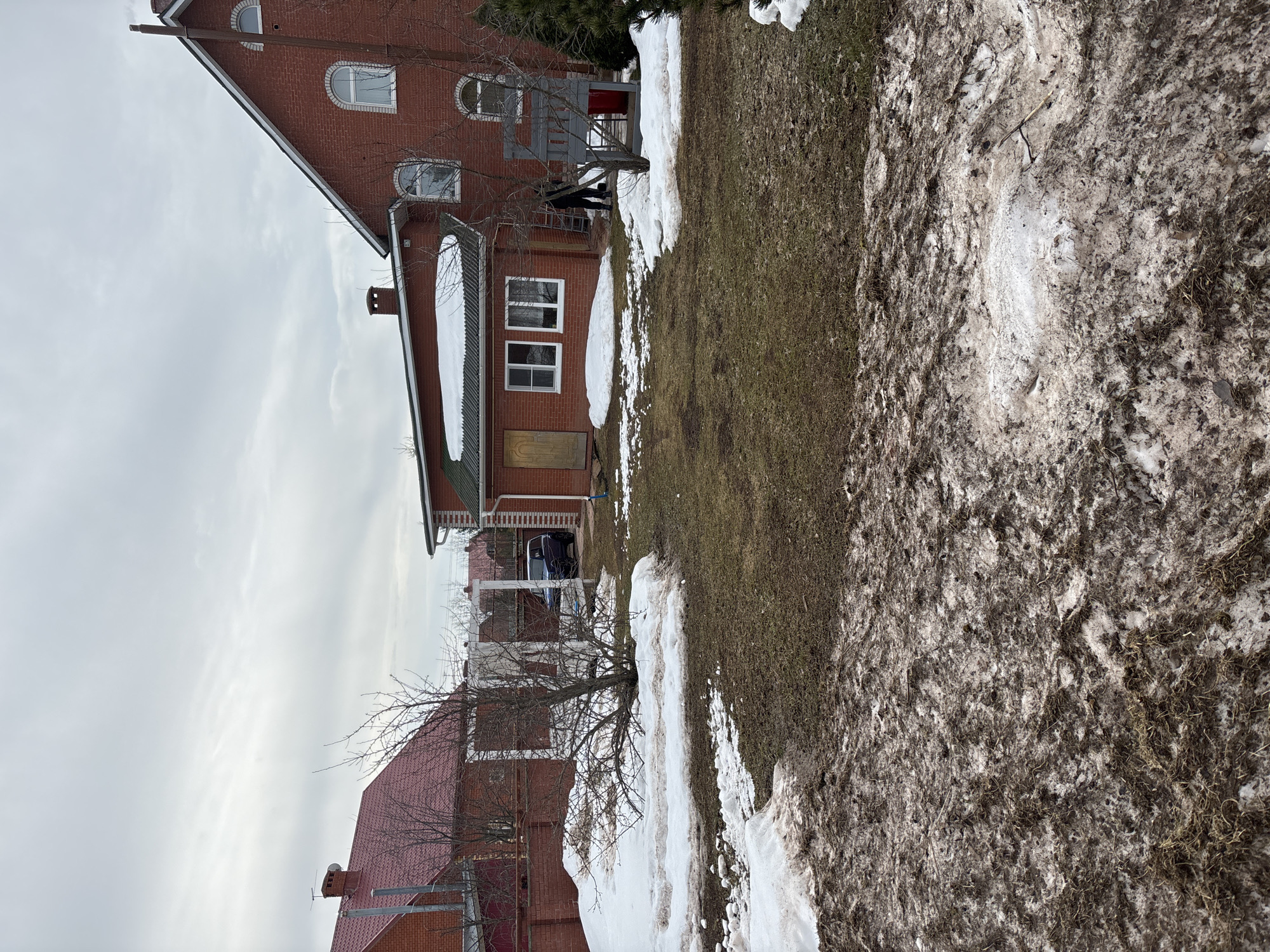 The image depicts a landscape on an overcast day in the Russian city of Ферма. In the foreground, a strip of dirty snow transitions into a grassy patch. Beyond this, two brick buildings stand, exhibiting classic Russian architecture with prominent gabled roofs and evenly spaced windows. A leafless tree is visible between the buildings. A car is parked between the buildings. The sky is overcast and grey.