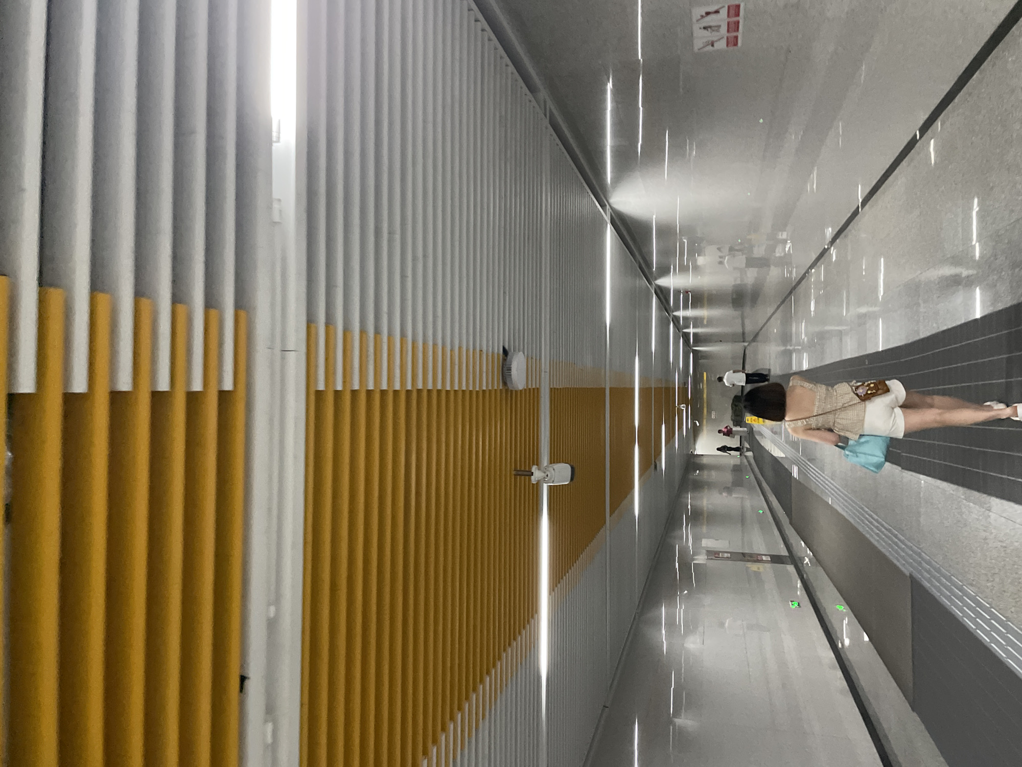 The image depicts a woman walking through a modern, well-lit underground corridor, likely a subway station or transit tunnel, in Liangjiang, China. The ceiling is a prominent feature, composed of horizontal louvers that transition from a bright yellow on the left to a light gray on the right. Fluorescent lights are embedded within the gray section of the ceiling, illuminating the space.
The woman in the foreground is walking away from the camera, towards the gray section of the ceiling. She is wearing a beige patterned sleeveless top and white shorts. She carries a light blue tote bag over her left shoulder and a small brown bag with a floral design on her right hip. Her hair is dark and cut to shoulder length.
The corridor itself is long and appears to be a moving walkway or a wide pedestrian path. The walls are smooth and light-colored, with several green illuminated markers spaced along the left wall. Reflections of the ceiling lights are visible on the polished floor. In the distance, other individuals can be seen walking in both directions. To the right of the corridor, there is a darker strip, and beyond that, a wall with some signage. On the left side, there's a dark gray area, possibly a non-moving section of the walkway. A security camera is visible mounted on the ceiling in the yellow-to-gray transition zone. The overall impression is one of a clean, functional, and contemporary urban transit space.