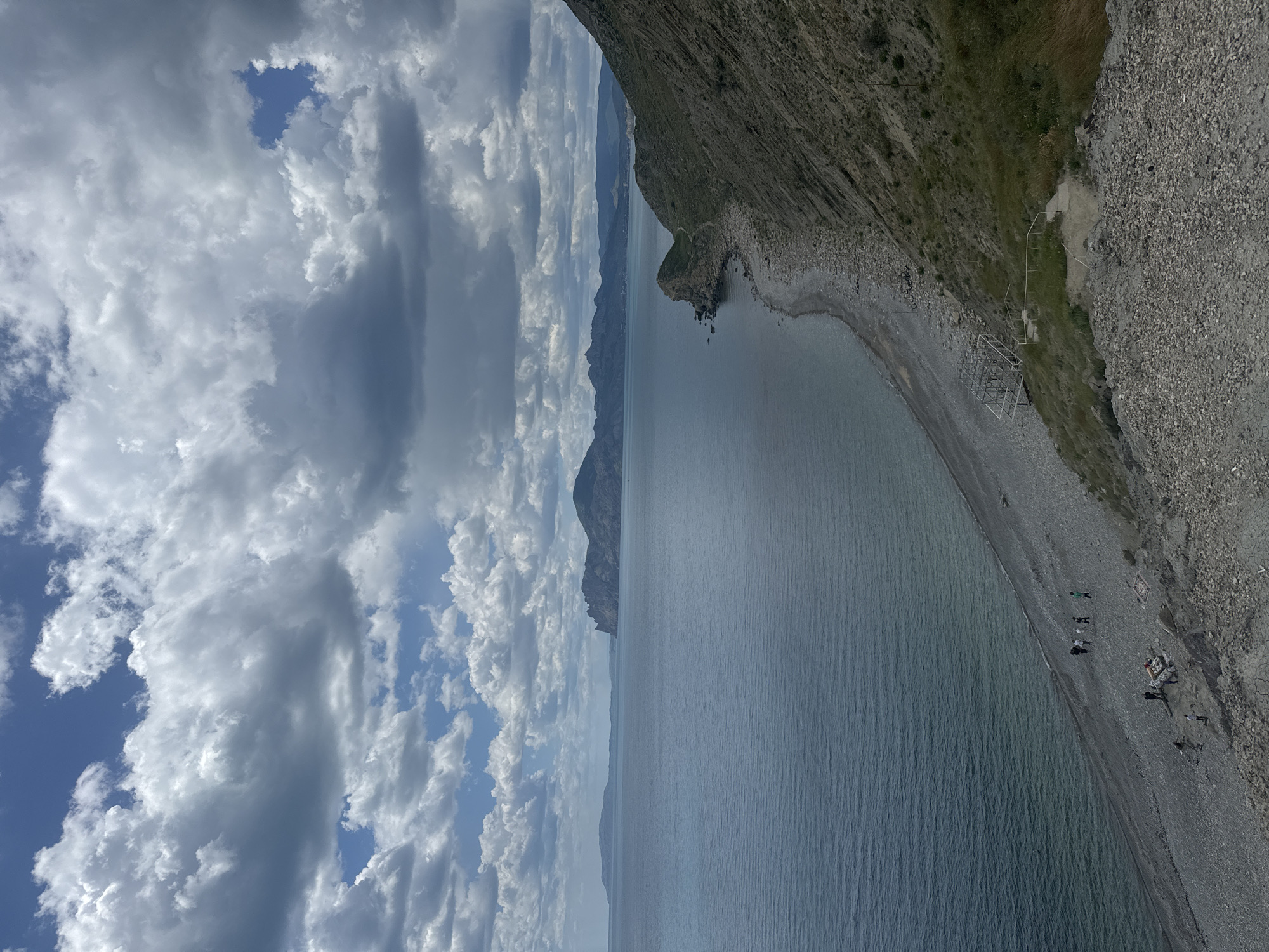 The image shows a coastal scene in Орджоникидзевский поселковый совет, Ukraine. The sky is partially covered with white and gray clouds. The sea is calm and blue-gray. A rocky cliff dominates the right side of the image, sloping down to a pebble beach. Some people are on the beach. A metal structure is located on the beach near the cliff. Mountains are visible in the distance, partially obscured by haze.