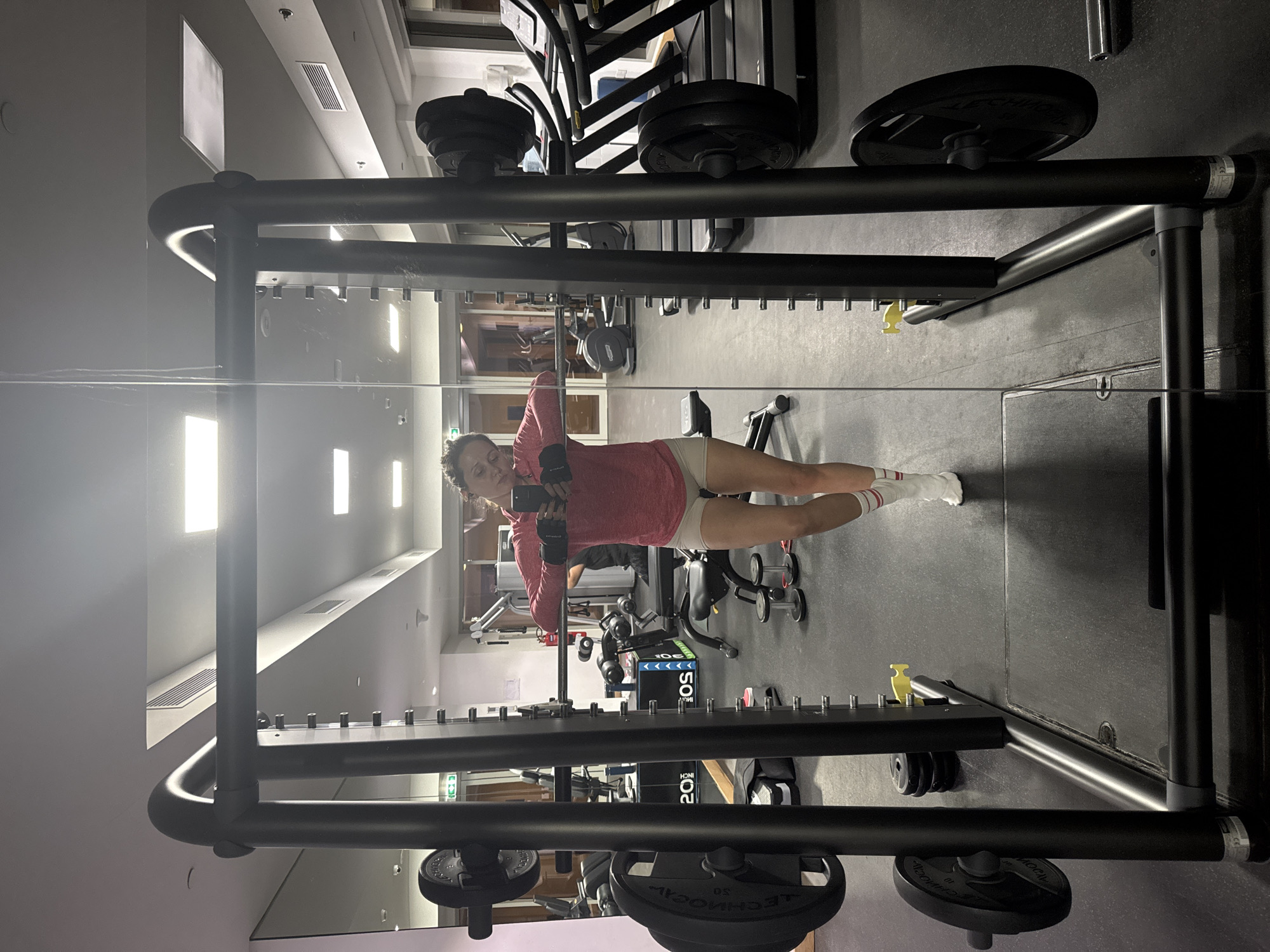 The image shows a woman in a gym setting, possibly in Dubai, United Arab Emirates, given the presence of fitness equipment typically found in such facilities. The woman is standing in front of a mirror, taking a selfie with her phone. She is wearing a red hoodie, light-colored shorts, and striped socks. The gym appears well-equipped, featuring weight machines and free weights. The floor is a dark gray or black color. The presence of windows and interior lighting suggests an indoor location.