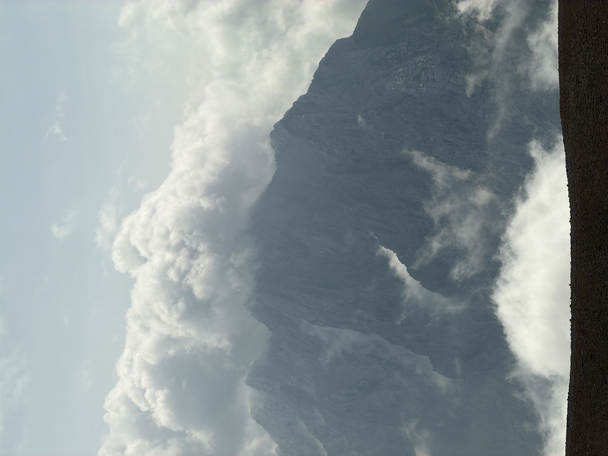 A majestic mountain range, shrouded in wispy clouds, dominates the frame. The peaks, etched with rugged texture, rise sharply against a pale, overcast sky. Patches of white fog swirl around the mountainsides, partially obscuring their formidable presence. In the foreground, a dark, stony slope stretches across the bottom of the image, providing a stark contrast to the ethereal landscape above. The overall impression is one of raw, natural beauty, evoking a sense of awe and solitude. This scene could be located in the Bavarian Alps, near the picturesque town of Garmisch-Partenkirchen, Germany, a region renowned for its dramatic mountain scenery.