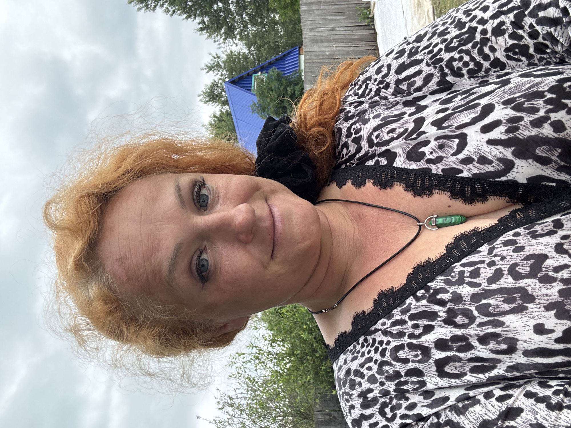 An outdoor, medium shot selfie shows a white woman with red curly hair and blue eyes. She is wearing a black and white leopard print top with black lace trim. A dark cord necklace with a green pendant hangs from her neck. She is positioned in front of a wooden fence, trees, and a house with a blue roof. The weather appears overcast. The location is identified as Chaikovsky, Russia.