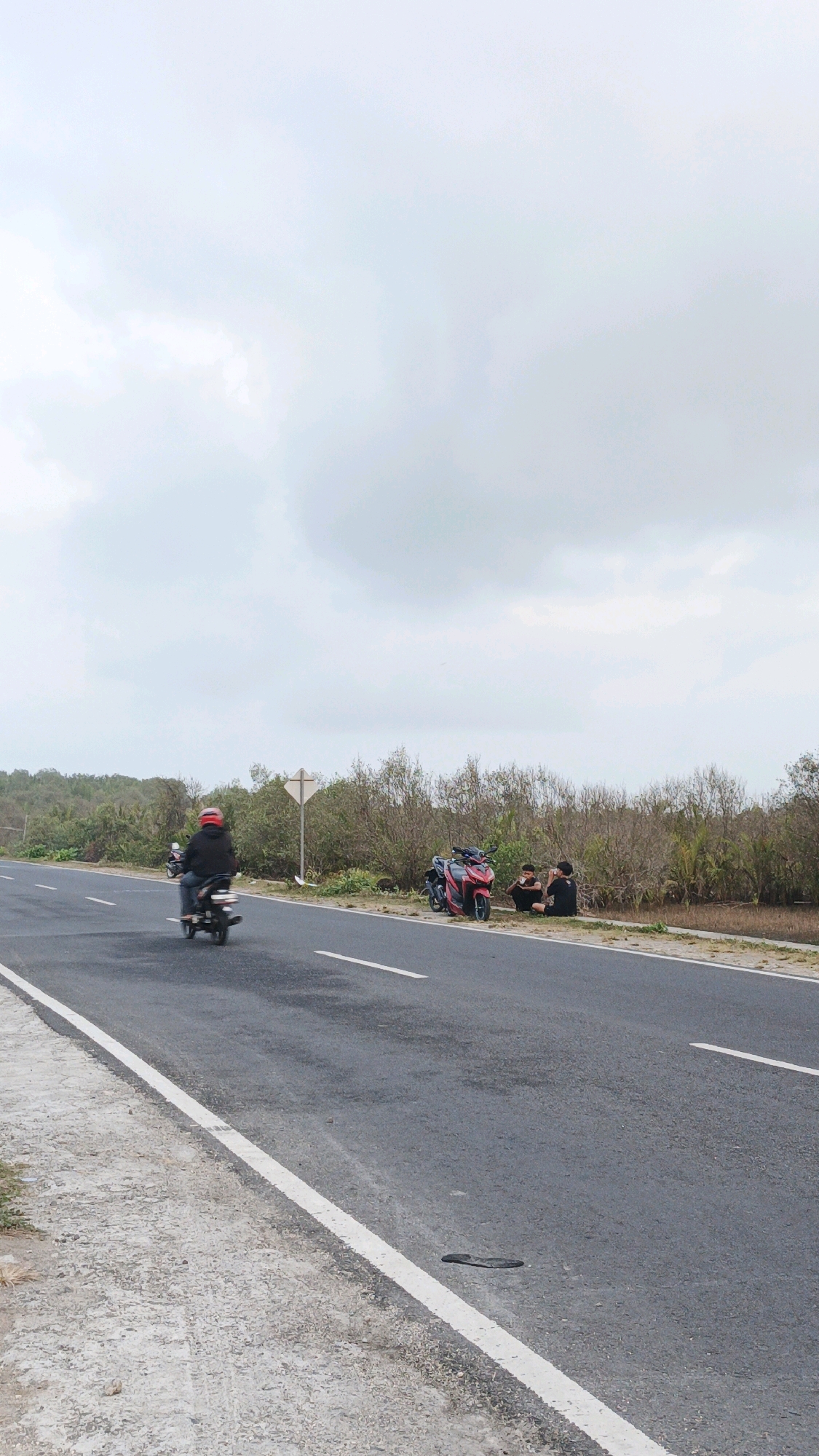 The image depicts a roadside scene in Pangandaran, Indonesia. On the asphalt road, a person wearing a red helmet is riding a dark-colored motorcycle. The road has white lane markings. To the right of the road, a group of people are sitting near two motorcycles, one red and one black. The environment is characterized by sparse vegetation and a cloudy sky. The overall impression is that of a rural or coastal road.