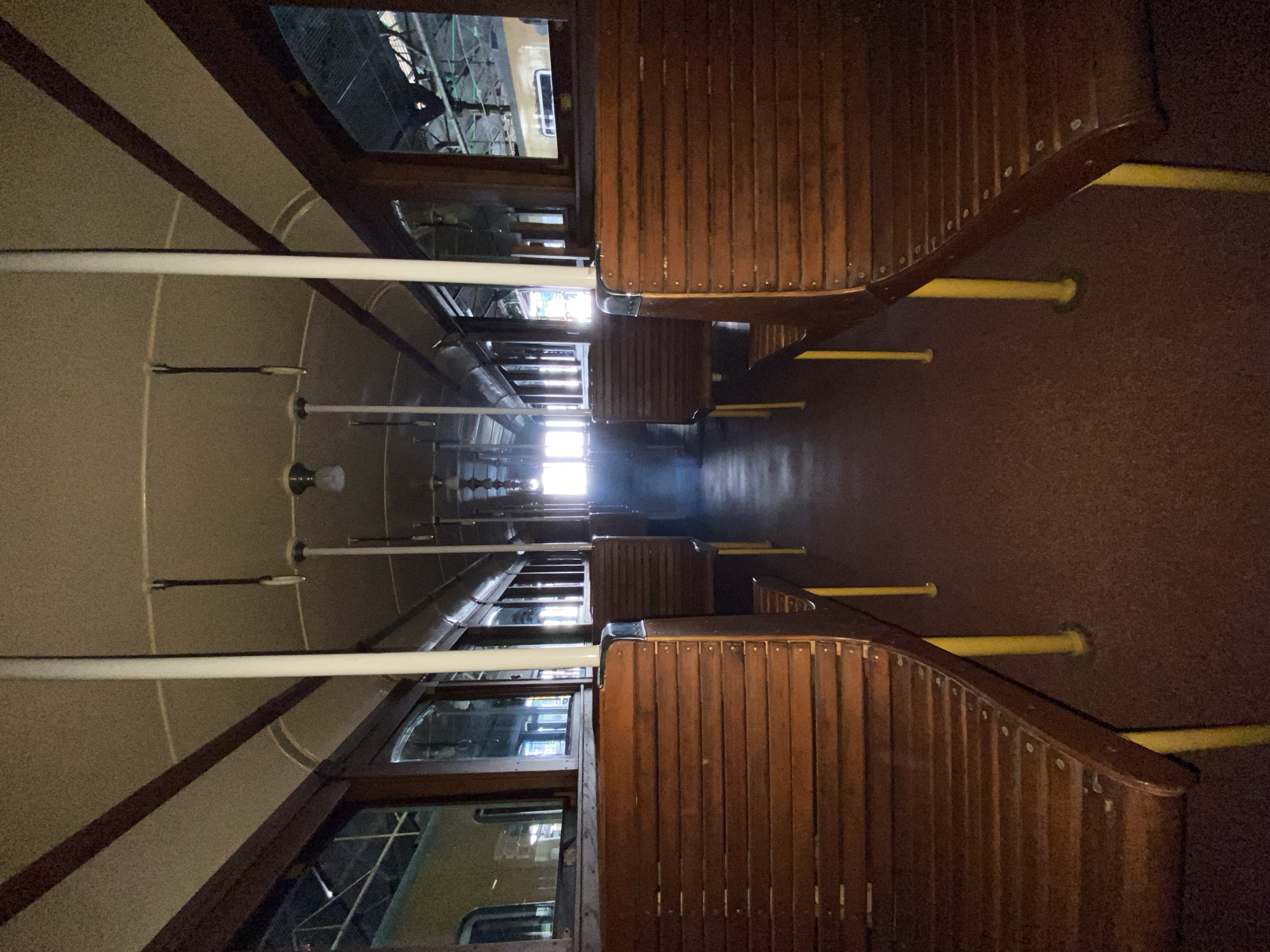 The image shows the interior of a vintage Buenos Aires subway car. Rows of dark wooden seats line both sides of the car. Yellow poles, presumably for support, extend from the seat backs toward the floor. Overhead, white grab bars run the length of the car. Windows reveal glimpses of what appears to be a maintenance facility or another subway car. Bright light illuminates the end of the car. The location is likely within the Buenos Aires, Argentina subway system (Subte), known for preserving historical aspects of its fleet.