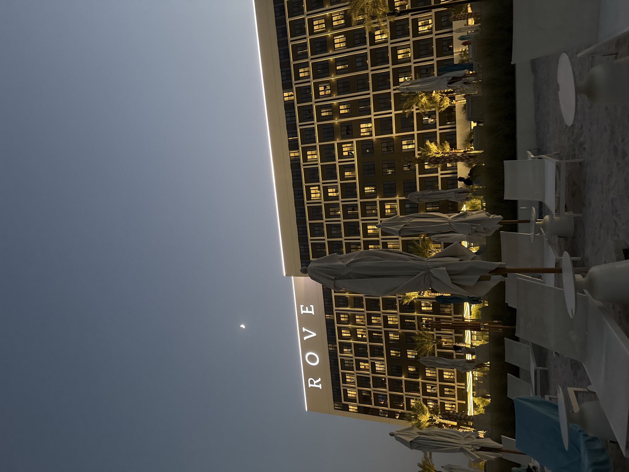 The image depicts a beachfront hotel in the city of Unknown, United Arab Emirates, at dusk. The hotel, branded with illuminated "ROVE" signage, is a large, multi-story building with numerous lit windows. In the sky, a crescent moon is visible. In the foreground, rows of white lounge chairs and closed beach umbrellas are arranged on the sand. Palm trees and other greenery are situated between the beach and the hotel. The overall scene suggests a tranquil evening atmosphere at a resort.