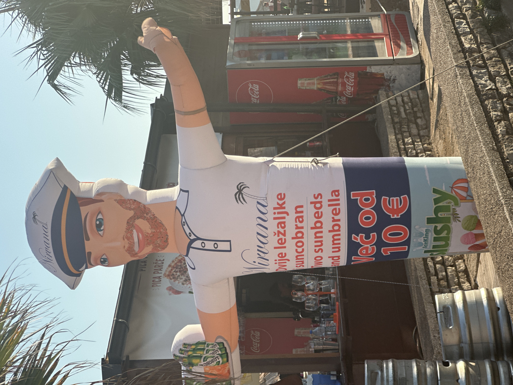The image captures a cheerful, inflatable mascot in a sailor's hat and polo shirt, positioned outdoors in front of a beach bar. The mascot prominently displays text in Croatian and English: "Nirvana," "Dvije ležaljke suncobran" (Two sunbeds and umbrella), and "Već od 10 €" (From 10 €), along with "IceKing Slushy" and images of ice cream cones. The bar itself has a Coca-Cola refrigerator, bottles of beverages, and glasses on display. Palm trees frame the scene, suggesting a coastal location. The mascot's arms are outstretched, one holding a tray of beer bottles. The ground is paved with stone and there are metal kegs stacked to the left. The overall ambiance is relaxed and inviting, typical of a vacation spot. The location is the beach bar "Nirvana" in Rafailovići, Montenegro, a popular tourist destination known for its beaches and waterfront amenities.