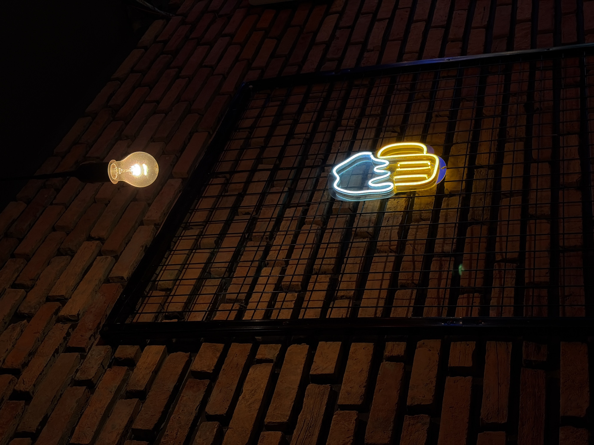 The image shows a neon sign shaped like a beer mug hanging on a brick wall. The neon sign is blue and yellow. A bare lightbulb hangs from the ceiling above the beer mug sign. The brick wall is rough and weathered. A metal grate is in front of the beer mug sign. The lighting is dim, with the neon sign and lightbulb being the primary light sources. The scene appears to be indoors, possibly in a bar or restaurant. The image is taken from a low angle, looking up at the neon sign.
