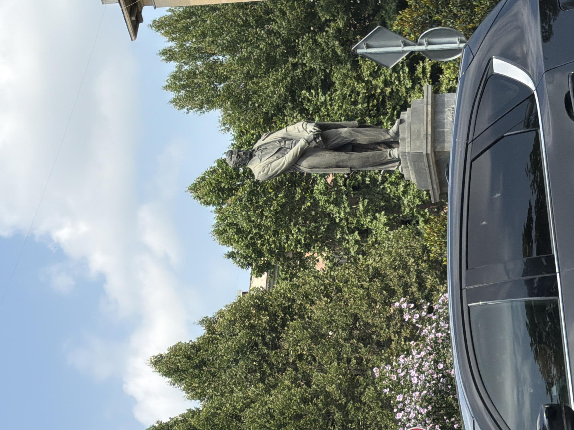 The image shows a bronze statue of a man in a suit and tie, standing on a stone pedestal. The statue is positioned in front of a backdrop of lush green trees and a partially visible building with a slanted roof. A black car is parked in the foreground, obscuring the lower portion of the statue and pedestal. A traffic sign with a diamond-shaped sign and a circular sign is visible to the right of the statue. The sky above is blue with scattered white clouds. This statue is located in Freedom Square in Tbilisi, Georgia, and it depicts Zakaria Paliashvili, a prominent Georgian composer.