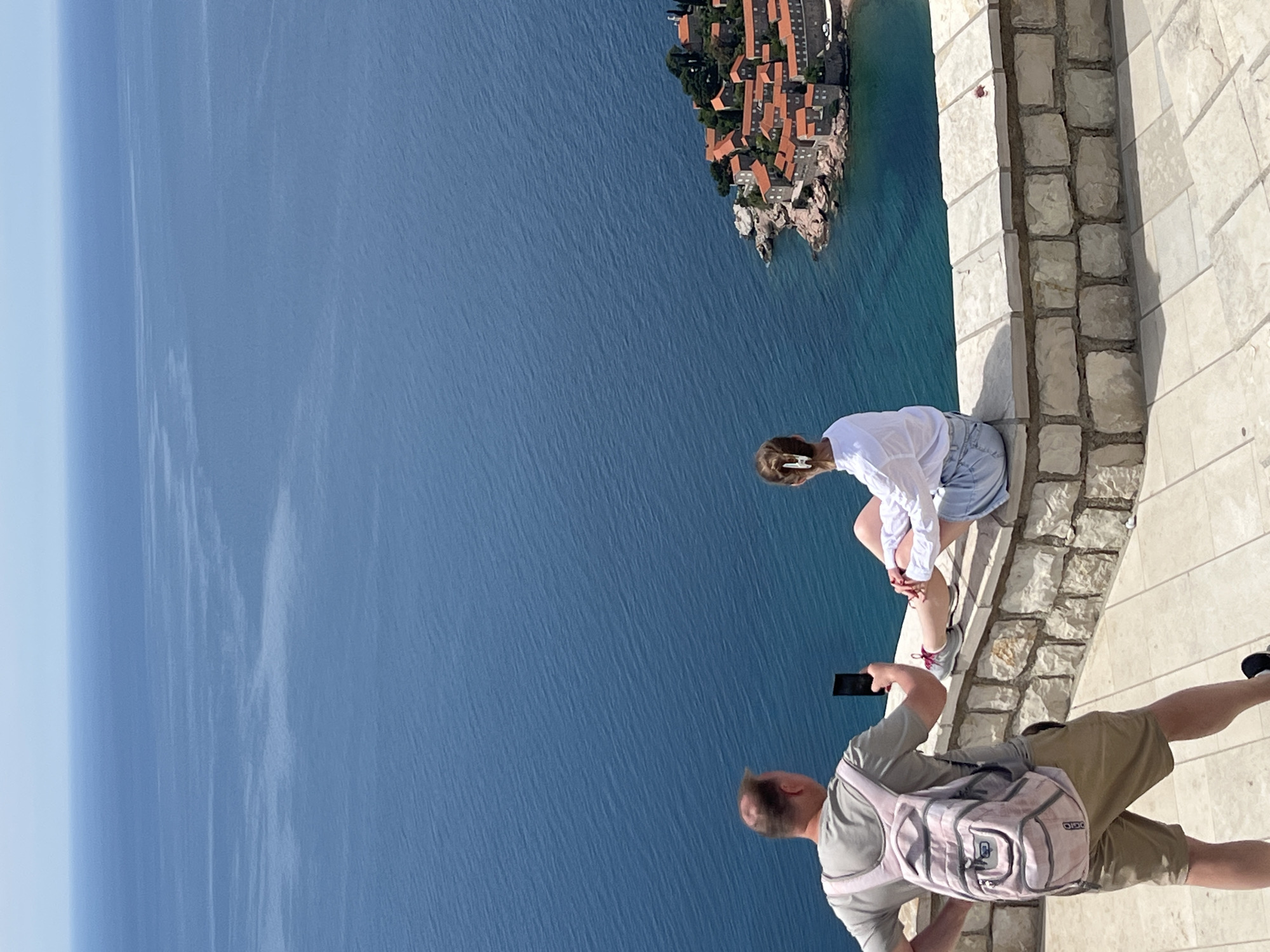 The image captures a scene in Sveti Stefan, Montenegro. From an elevated vantage point, a woman is sitting on a stone ledge near the edge. She is wearing a white shirt and a light-colored skirt or shorts. A man, with a backpack and a smartphone, stands nearby, seemingly taking a picture of her. The background shows the sea and an islet with buildings, likely part of the Sveti Stefan resort. The perspective is slightly tilted, giving a sense of height.