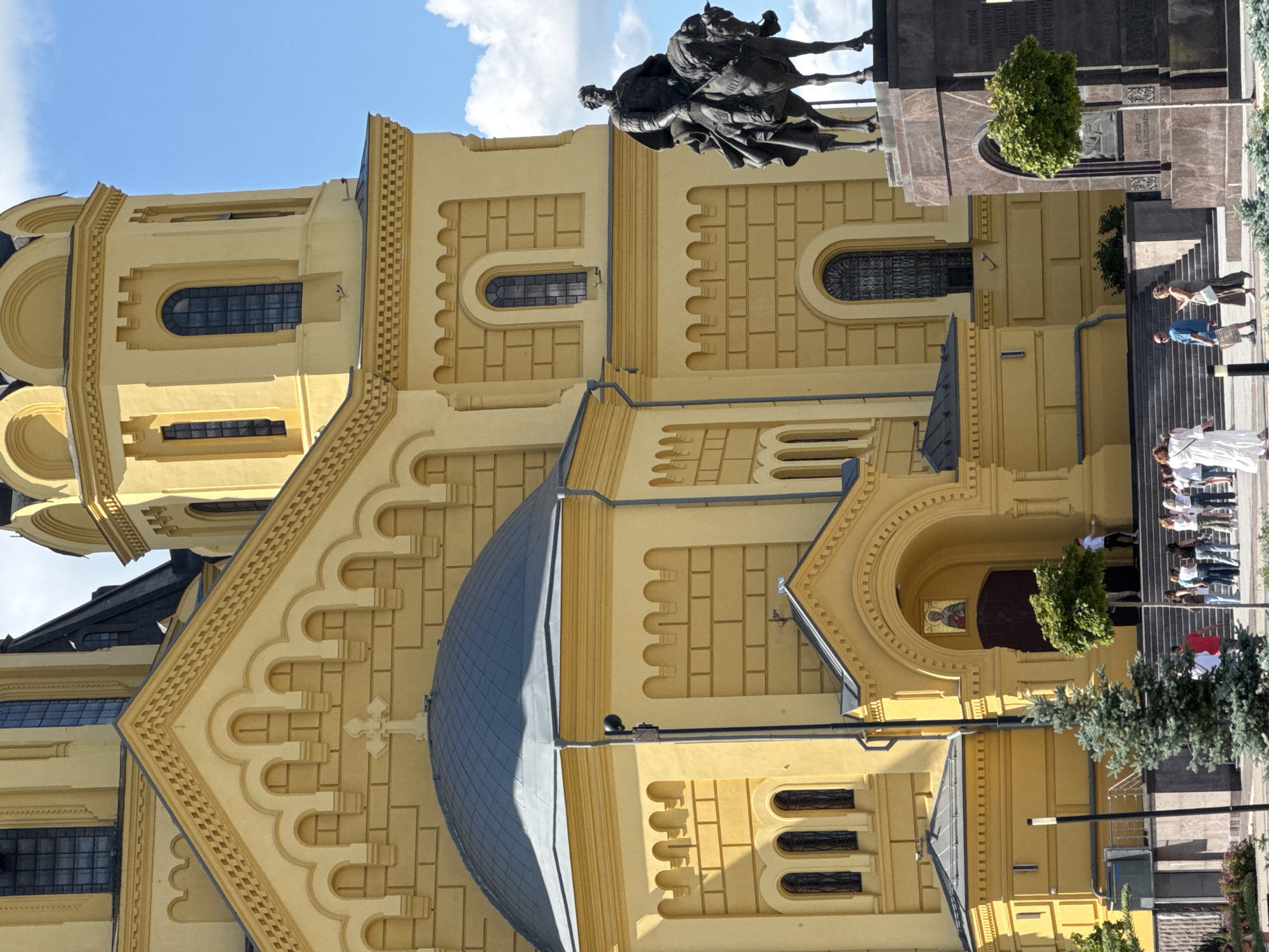 The image shows the Annunciation Cathedral in Nizhny Novgorod, Russia, on a sunny day. The cathedral is a large, yellow building with a blue dome and intricate architectural details. In front of the cathedral, there is a bronze equestrian statue of a man on a horse, positioned on a marble pedestal. A crowd of people, dressed in casual attire, are seen walking up and down the steps leading to the cathedral entrance. Some are looking at their phones, while others are talking to each other. A few trees are also visible in the foreground and around the statue. The sky is blue with scattered white clouds.