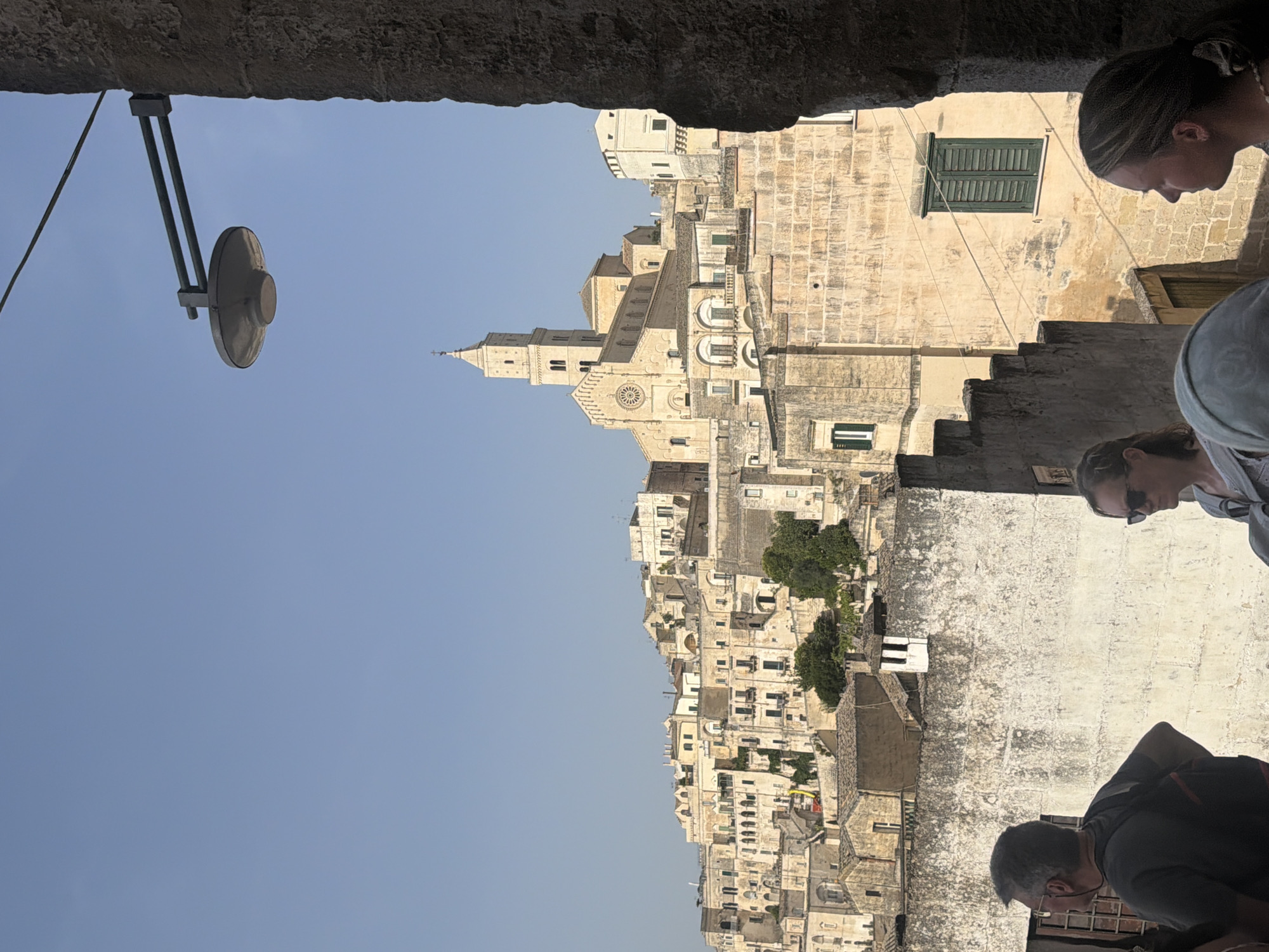 The image depicts a view of the Sassi di Matera, a cave dwelling district in Matera, Italy. In the foreground, the shoulders and backs of three individuals are visible. A man with grey hair and glasses is on the left, looking away from the camera. To his right, a woman wearing sunglasses faces away from the camera. Another woman is partially visible on the right edge of the frame, facing left.
The midground features a wall with a distressed texture, and beyond that, a dense urban landscape characterized by buildings constructed from pale stone. Many of these structures exhibit a multi-tiered arrangement, clinging to the hillside. A prominent bell tower with a cathedral-like structure rises above the buildings in the center-background, identifiable as the Cathedral of Matera. A street lamp is mounted on a stone column on the right side of the image, in the upper midground. The sky is clear and blue.