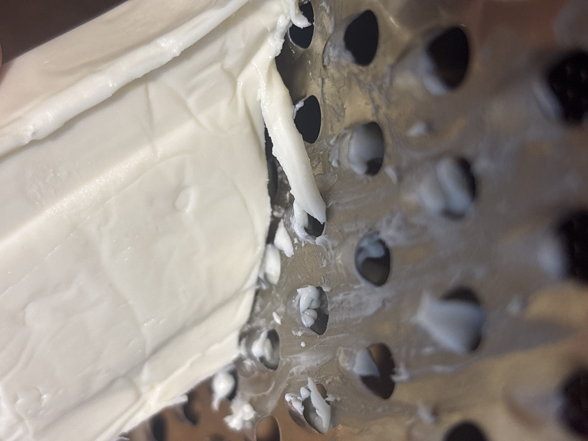 A close-up shot shows a block of white cheese being grated on a metal grater. Small shreds of cheese are visible on the grater and sticking to the cheese block. The grater has circular holes. The lighting is overhead, creating highlights and shadows. The background is dark and out of focus, making the cheese and grater the sole subjects. The image was taken in Kremenchuk, Ukraine.