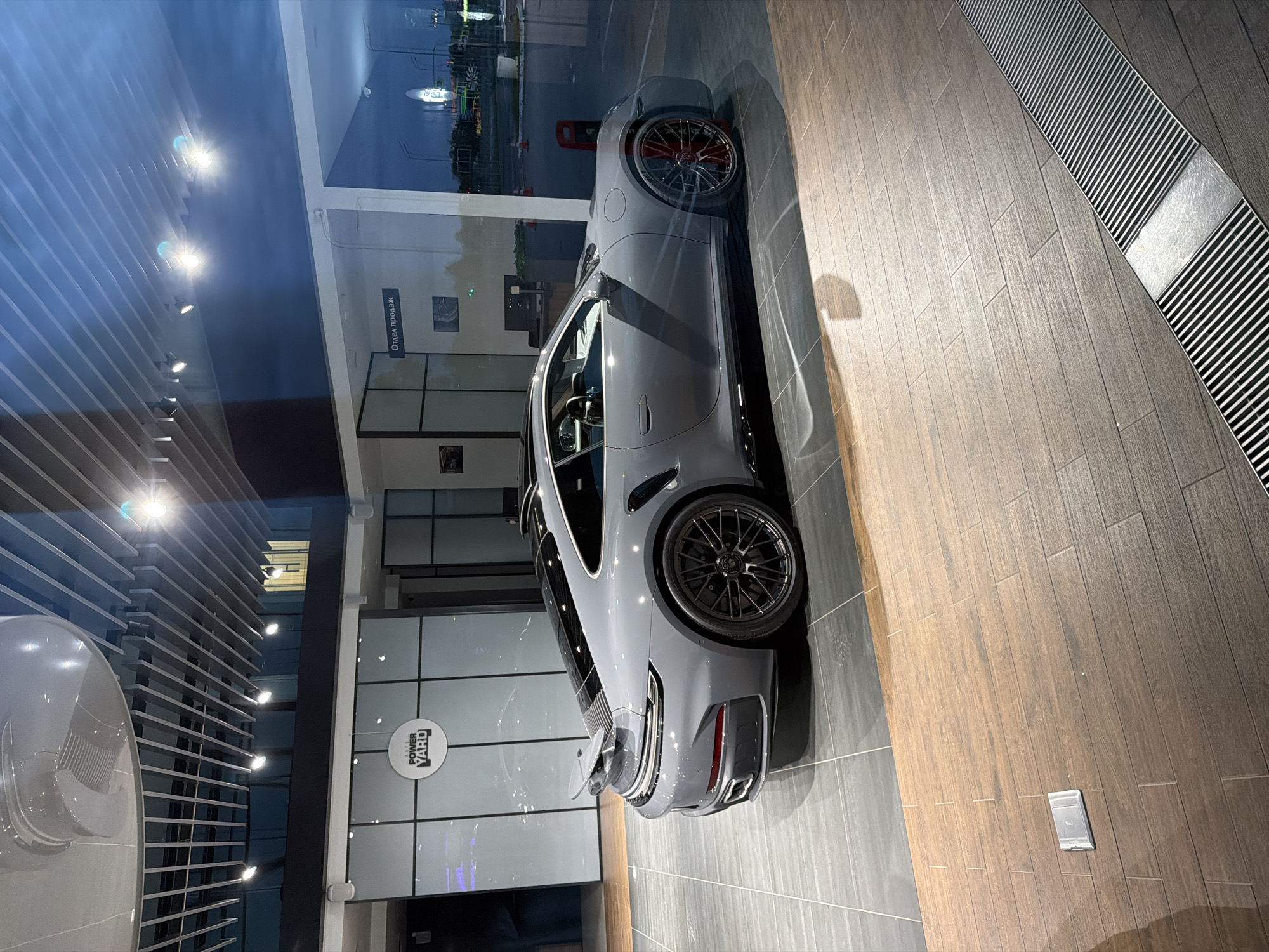A grey Porsche 911 Turbo S is parked inside a brightly lit, modern showroom with a reflective glass facade. The car is viewed from its rear three-quarter angle. It has a prominent rear spoiler, black multi-spoke wheels, and red brake calipers. The showroom interior features a tiled floor, a suspended ceiling with diagonal slats and spotlights, and frosted glass panels with a circular logo that reads "THE POWER YARD". Through the glass facade, a street scene at dusk is visible, with traffic lights and building lights. The floor is a mix of dark grey rectangular tiles and light brown wood-look planks. A silver electrical outlet is visible on the wood-look floor in the foreground.
