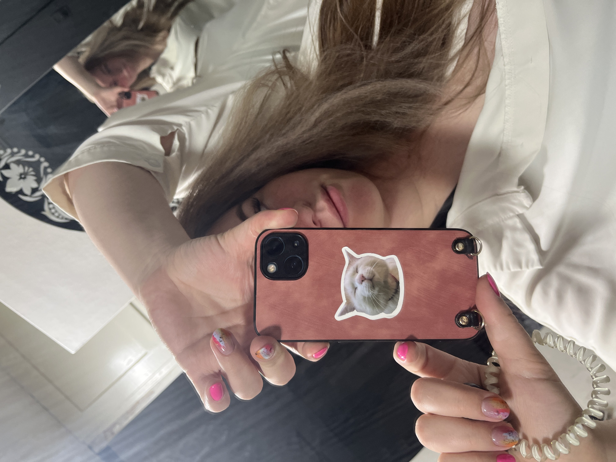 A person is taking a selfie in a mirror, holding a smartphone with a pink case and a cat sticker on the back. The person has brown hair and is wearing a white shirt. The nails on their left hand are painted with pink polish, while the nails on their right hand have an ombre design. A white spiral bracelet adorns their right wrist. The reflection in the mirror shows the person's face and the room they are in, which includes a black and white patterned surface. There is no information suggesting the photo was taken in the city of Chaikovsky, Russia.
