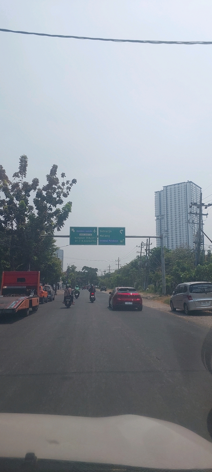 This image captures a street scene in Surabaya, Indonesia, under a hazy, overcast sky. A green overhead road sign indicates directions to "Bandara Juanda" (Juanda Airport), "Sidoarjo," "Malang," "Rungkut Industri" (Rungkut Industrial area), "Terminal Purabaya," and "Dr. Ir. H. Soekarno."
The road is busy with traffic, featuring several motorcycles, cars, and a flatbed truck. A red car is prominently in the foreground, partially obscuring a silver minivan. A tall, modern apartment building dominates the skyline on the right, with utility poles and lush green trees lining the street. The perspective suggests the photo was taken from inside a vehicle, looking forward. The overall impression is of a typical urban environment during daylight hours, possibly experiencing mild air pollution or humidity.