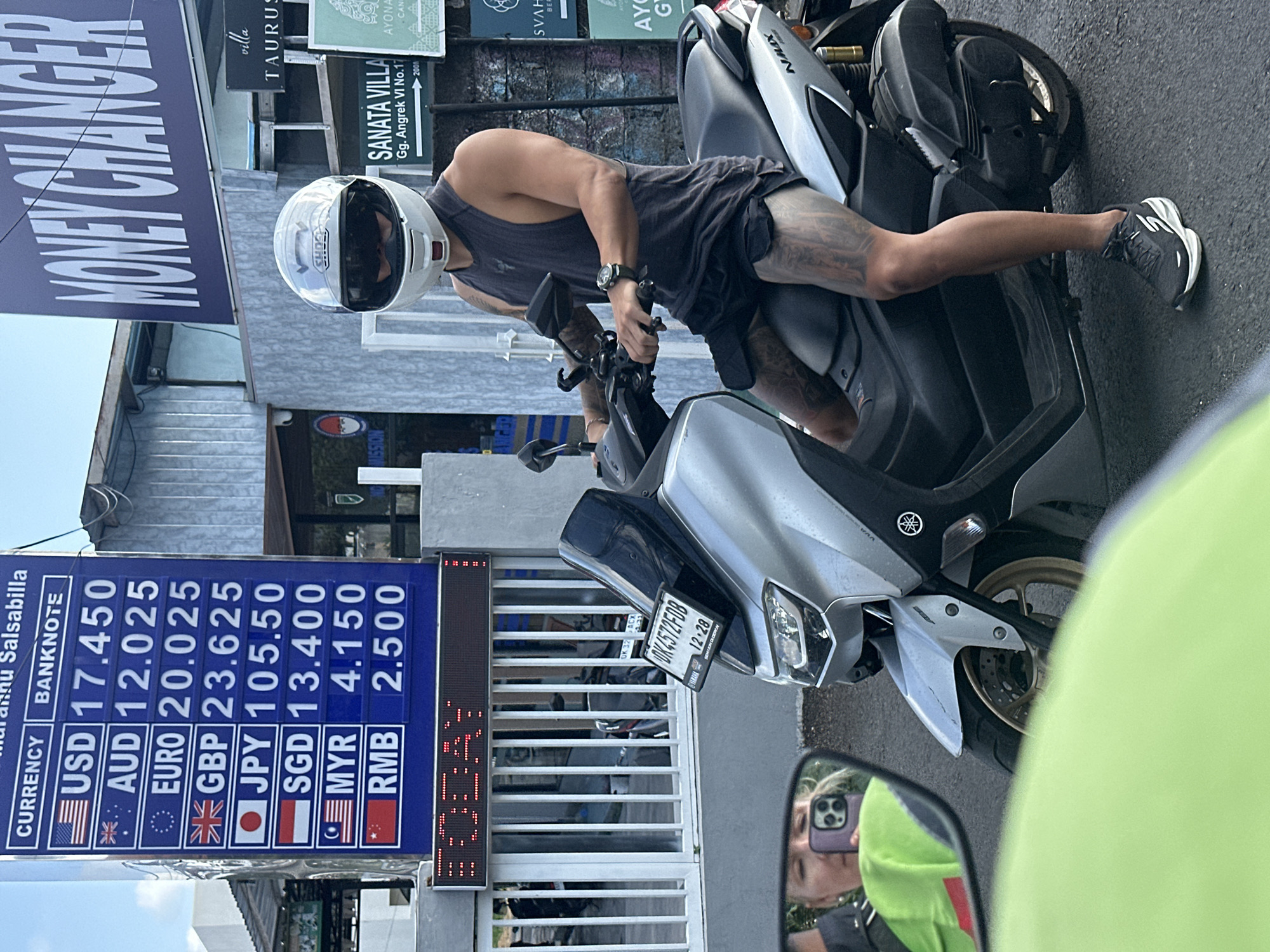 A man wearing a white full-face helmet, dark tank top, and black shorts sits on a silver Yamaha NMAX scooter. He has extensive tattoos on his legs. The scooter's license plate reads "DK4572FDB". In the background, a blue and white currency exchange rate board displays exchange rates for various currencies including USD, AUD, EURO, GBP, JPY, SGD, MYR, and RMB, with numerical values next to each currency. Above the currency board, a blue sign reads "MONEY CHANGER". To the right of the currency board, a sign for "SANATA VILLA Gg. Angrek VI No.17" is visible. The environment appears to be outdoors, with a paved road and a building structure. A reflection in the scooter's rearview mirror shows a person holding a smartphone and wearing a bright green vest. The scene is located in North Kuta, Indonesia, as indicated by the "DK" license plate prefix, commonly used in Bali, and the presence of a money changer and villas.