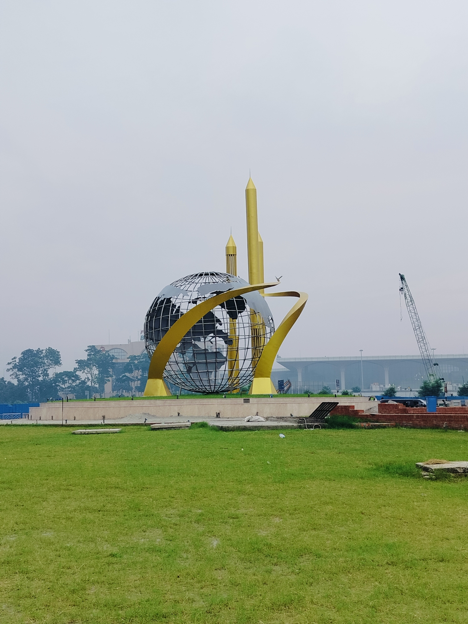 In Dhaka, Bangladesh, the iconic globe monument stands tall on a clear day, surrounded by lush green grass and a grey sky. The monument features a large, metallic globe with intricate details of continents, encircled by golden curved beams. Behind the globe, two tall, golden spires rise majestically, adding to the monument's grandeur. In the distance, a modern airport terminal with elevated walkways is visible, suggesting the monument's proximity to transportation hubs. A construction crane stands to the right, hinting at ongoing development or maintenance in the area. The scene is devoid of people, creating a serene and contemplative atmosphere.