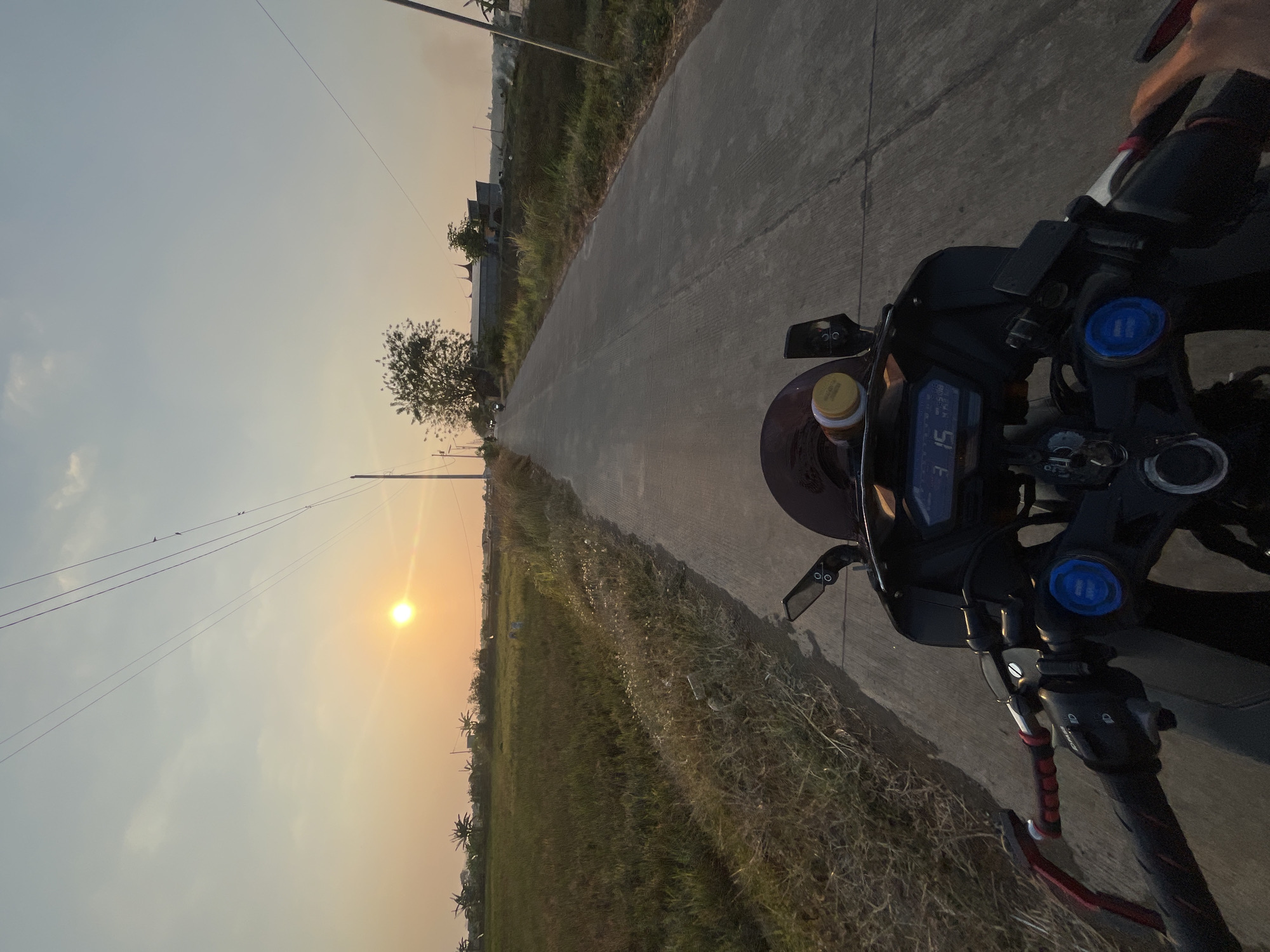 This is a first-person perspective, point-of-view shot from a motorcycle on a paved road in a rural area, likely in Pecangaan, Indonesia, during sunset. The sun, a bright orb, is low in the sky, casting a warm, golden light across the scene. To the left of the road, a field of dry, tall grass and some crops stretches towards the horizon. In the distance, there are scattered trees, utility poles with power lines, and a few buildings. The road itself is a two-lane paved surface with some visible cracks and power lines crossing above.
The motorcycle's handlebars and dashboard are prominent in the foreground, showing a digital display with numbers that appear to be '3 15'. The rider's hands are on the handlebars, with one hand visible reaching towards the right. The motorcycle is dark in color with some red accents on the handlebar grips. The overall atmosphere is serene and indicative of travel during the golden hour.
