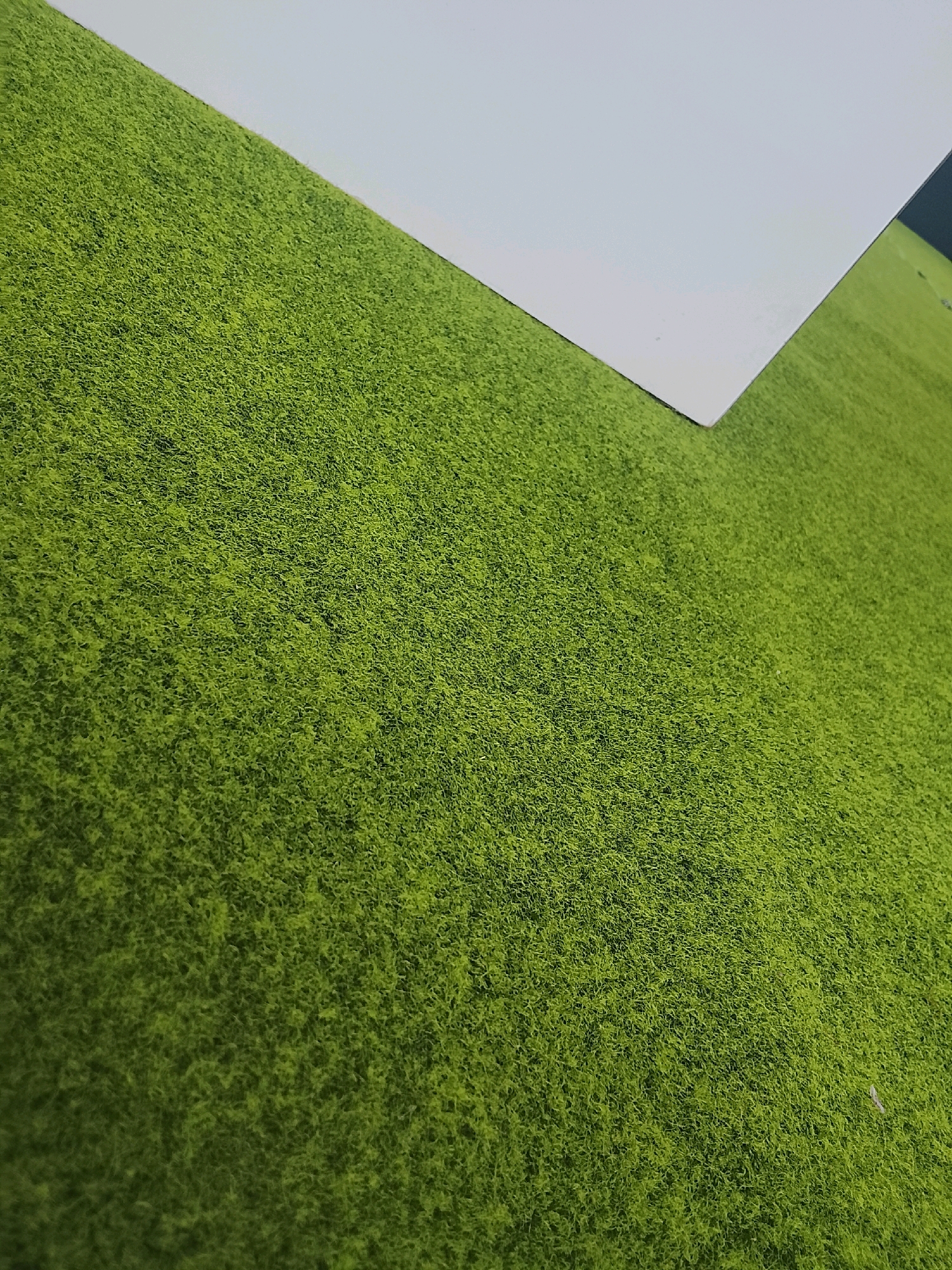 The image shows a close-up view of a textured green surface, likely artificial turf, with a white rectangular surface angled over it. The green surface appears to be on a flat plane, with the white surface casting a subtle shadow. The lighting suggests it is outdoors during the day, but no specific landmarks or environmental features that would pinpoint a location in Surabaya, Indonesia are visible. The image is a still shot with no people or actions depicted.