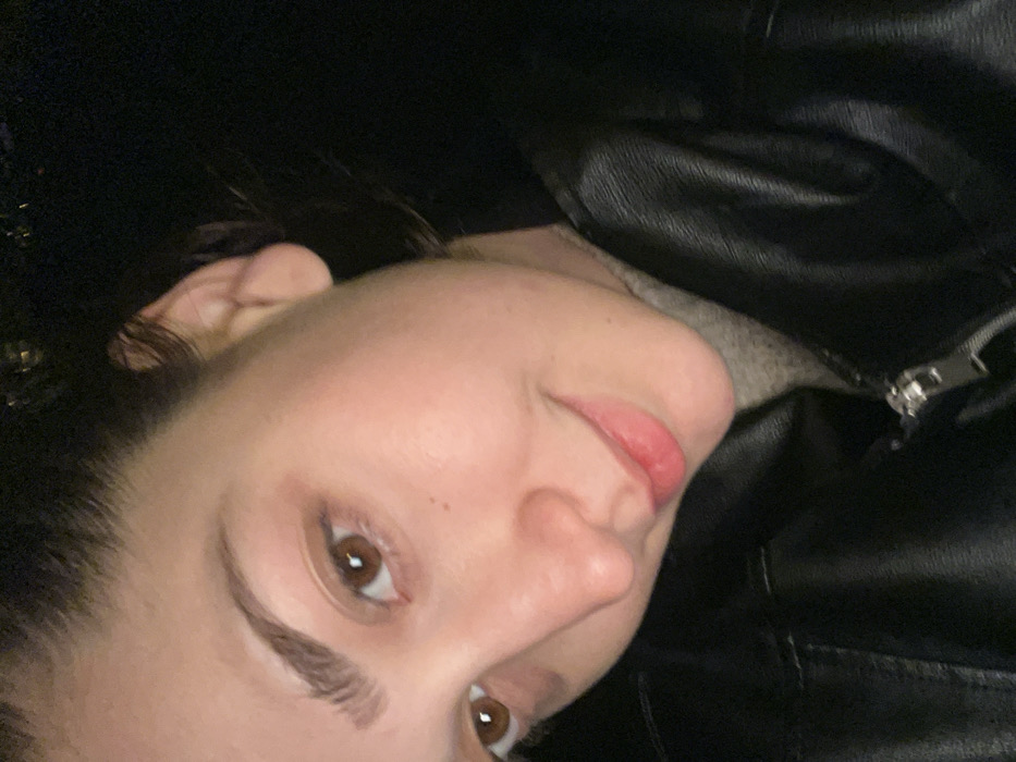 A woman's face is closely framed, angled slightly upwards. She has fair skin, dark hair, and brown eyes. Her lips are a soft pink. She wears a black leather jacket. A glimpse of a light grey sweater is visible beneath the jacket. The overall lighting suggests a dimly lit environment. A metal zipper is visible on the right side of the frame.