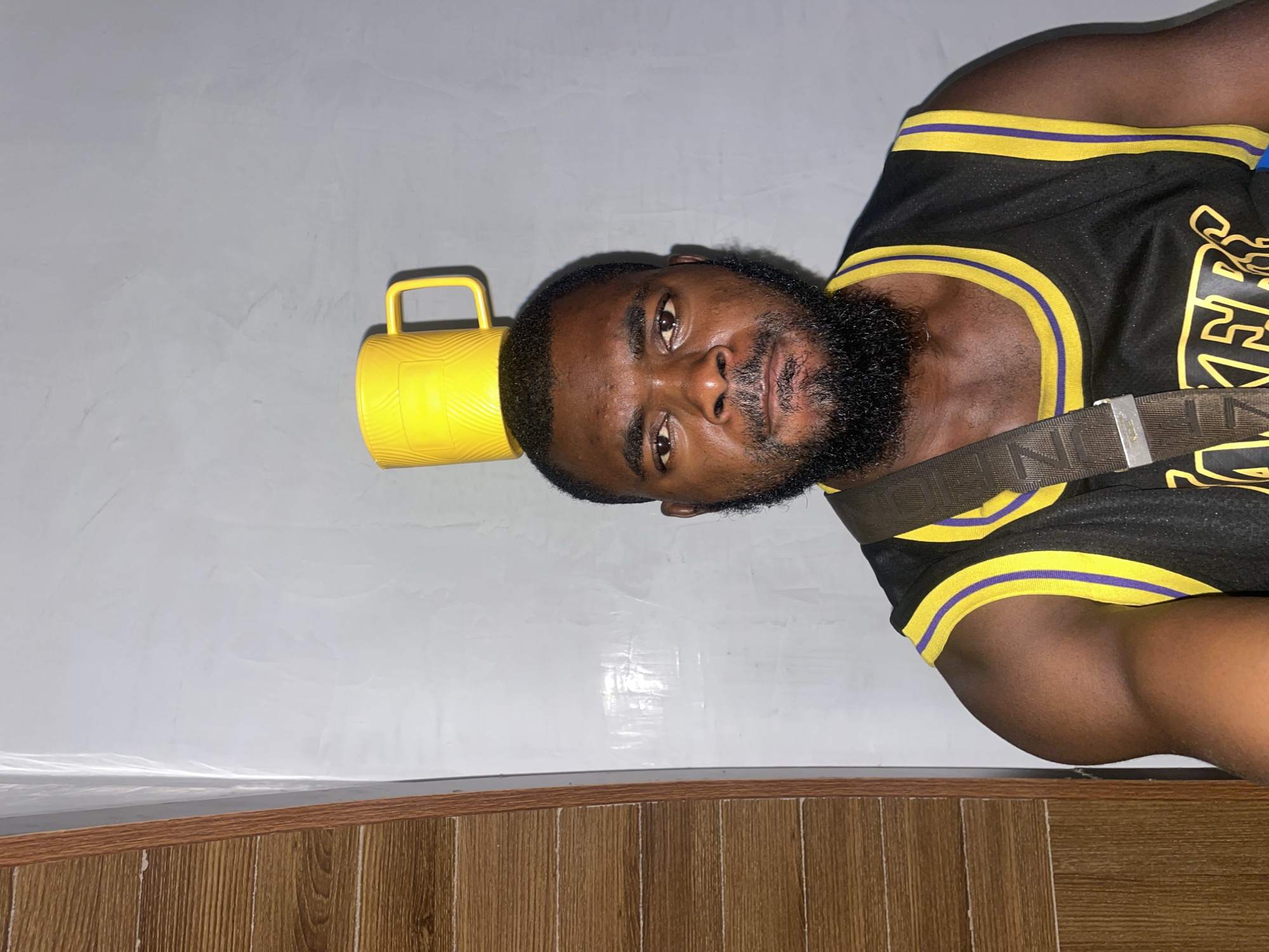 A close-up shot captures a dark-skinned man with a full beard wearing a black and gold basketball jersey. A yellow mug rests on his head. The background consists of a white wall with light variations. A wood-grained section is visible along the bottom of the frame. The location is possibly Jos, Nigeria, but this cannot be confirmed from the image. The man is the primary subject. There is no visible interaction or action occurring in the frame.