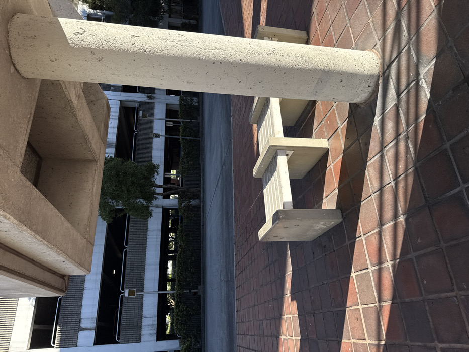 A low-angle shot shows a concrete architectural setting. A large, cylindrical concrete beam extends horizontally above. To the right, the beam casts a shadow on a red brick wall with protruding concrete steps. In the background, a parking structure is visible, with trees lining the side. The image is rotated sideways.