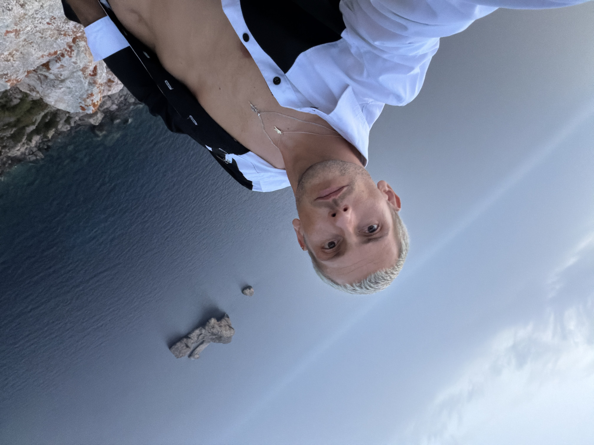 A close-up, high-angle selfie shows a person with short, bleached-blonde hair, a fair complexion, and stubble. They are wearing a partially unbuttoned white shirt with black accents and two silver necklaces. The person is positioned on a rocky cliff overlooking a dark blue body of water. In the distance, two small rock formations are visible in the water. The sky is overcast with wispy white clouds. The location is identified as near Sant Antoni de Portmany, Spain.