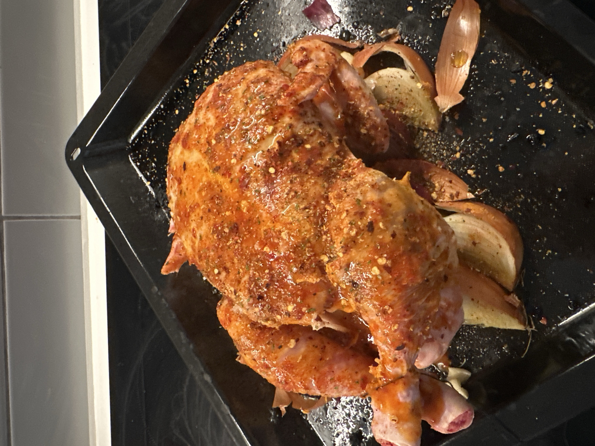 A seasoned and roasted whole chicken sits on a black baking pan, accompanied by onion slices. The pan rests on a stove top within a domestic kitchen setting. No people are visible. The image appears to be taken from above at a slight angle. There is no visible connection to the city of London, United Kingdom.