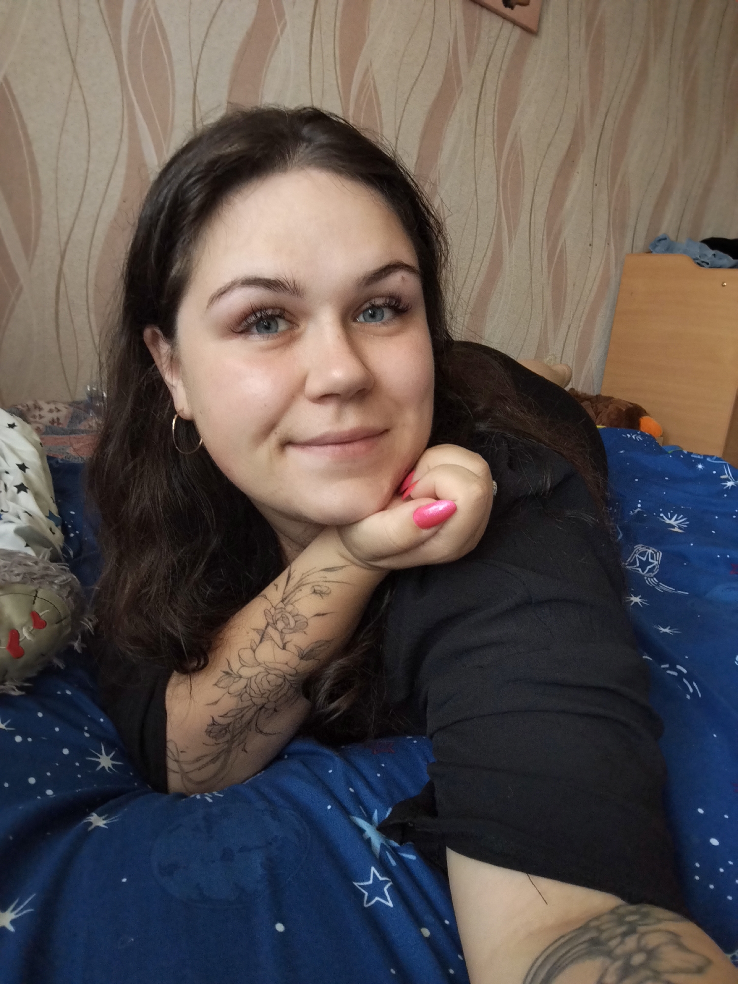 A woman with dark brown hair and blue eyes lies on a bed. She is wearing a black shirt and has tattoos on both forearms. Her nails are painted pink. The bedding is blue with a star and space pattern. There is a stuffed toy next to her. The background shows a wall with a light brown and cream striped wallpaper and a wooden cabinet or nightstand. The location is Chaikovsky, Russia.