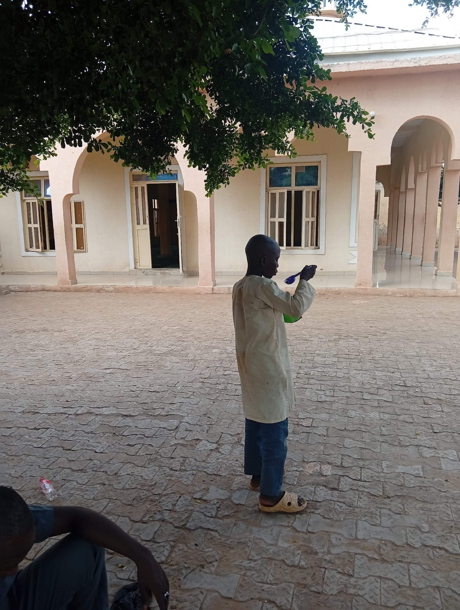 The image depicts a young boy, appearing to be of African descent, standing in an open courtyard. He is wearing a light-colored, long-sleeved tunic and blue trousers, along with light-colored sandals. The boy is holding a blue utensil in his right hand and a green bowl in his left, suggesting he might be eating or preparing to eat. Behind him, a building with a pale pink facade features arched openings and several windows with white frames. A large tree with dark green foliage is prominent in the upper left portion of the image, casting shadows. The ground of the courtyard is paved with interlocking stone tiles. In the lower left corner, a person is partially visible, seated on the ground with their back to the camera, wearing dark clothing. A clear plastic bottle with a red cap is also present on the ground near the seated person. The overall setting appears to be an outdoor space associated with a building that resembles a mosque or similar community structure. This scene is located in Zongoma Kyari, Nigeria.