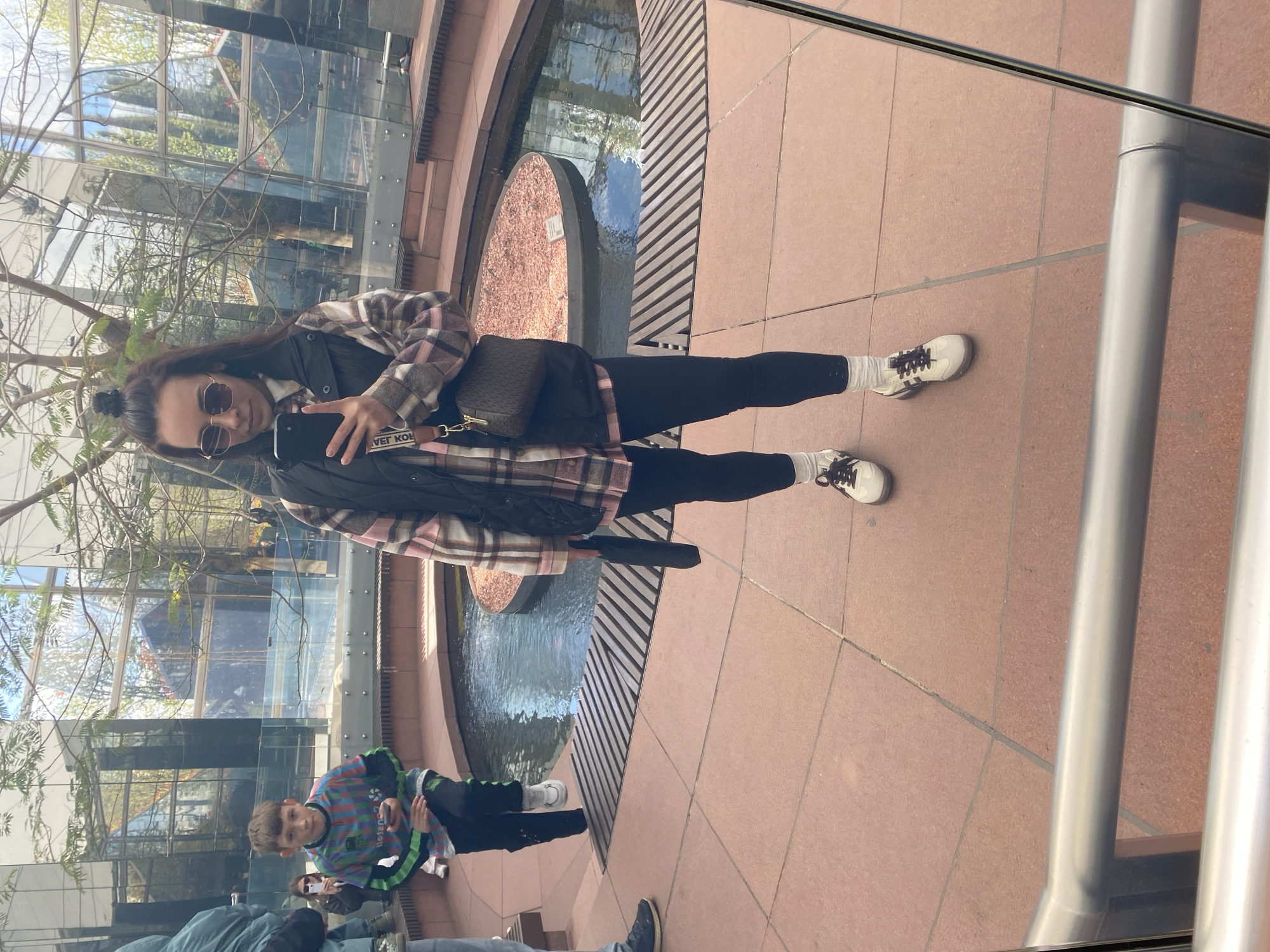 A person stands in front of a reflective glass surface, likely a window or mirror, taking a selfie with a phone. They wear sunglasses, a plaid shirt, a black jacket, leggings, and white shoes with dark laces. They have a small bag slung across their body. A shallow decorative pool or fountain is visible behind them. Reflections in the glass show additional people and the surrounding architecture, including trees and building structures. The reflection of one person is wearing a multi-colored shirt and is seated near the bottom left of the image. The environment appears to be an indoor atrium or walkway adjacent to an exterior space. The location is not determined to be Krasnodar, Russia.