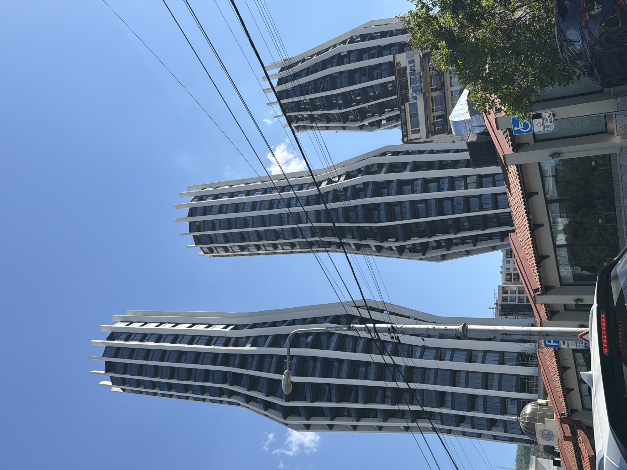 The image shows a view of three curved, modern high-rise buildings in Tbilisi, Georgia, as seen from street level. The buildings feature a white-and-dark-windowed facade. Power lines are strung across the sky. A street lamp and traffic signs are visible in the foreground. A sidewalk with a bus shelter and accessible parking is partially visible on the right side of the image. A tree and parts of vehicles are also present. The sky is clear blue with scattered clouds.