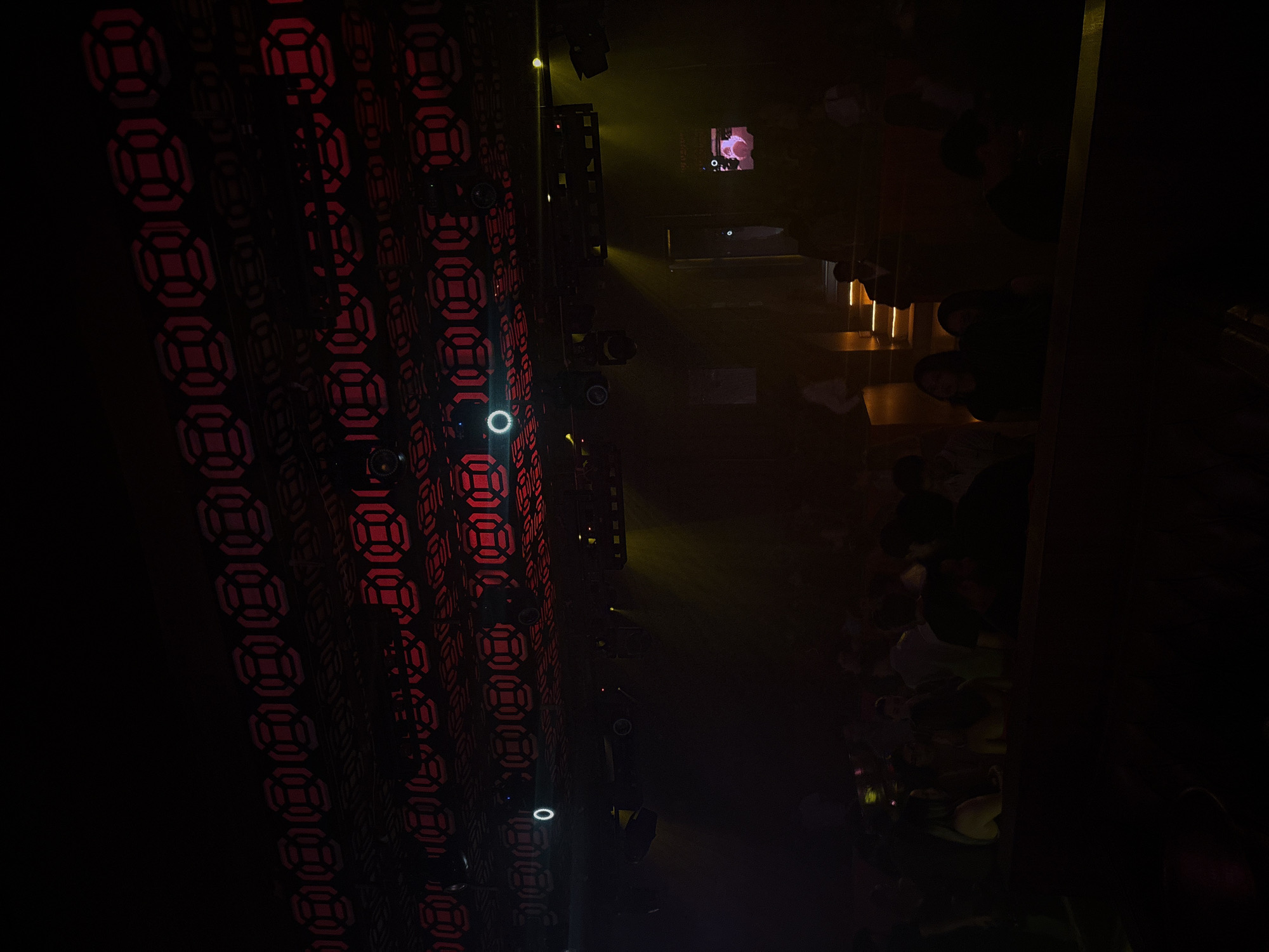 The image depicts a dimly lit interior, likely a club or entertainment venue in Puebla, Mexico. A patterned wall, illuminated with red geometric shapes, stretches along the left side of the frame. Stage lighting equipment is visible, projecting beams of yellow light into the space. A small screen displays what appears to be a video clip of a person, perhaps part of a performance. A crowd of people is gathered, some sitting at tables and others standing. The overall atmosphere suggests a social event or entertainment setting.