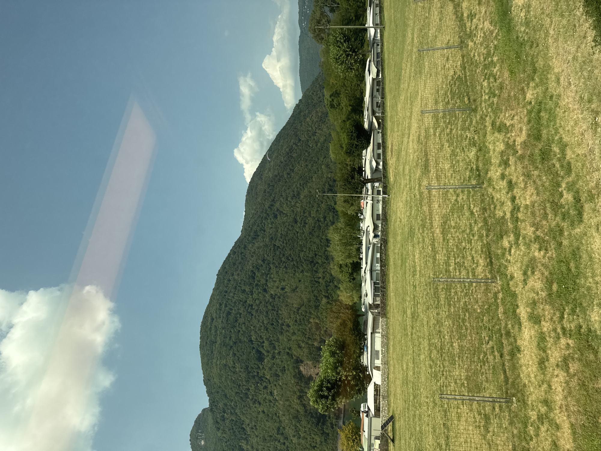 This is a landscape photograph taken in the city of Val Mara, Switzerland. The image captures a campsite situated at the base of a densely forested mountain under a clear blue sky with scattered white clouds. In the foreground, a grassy field is partially enclosed by a wire fence. Behind the field, a row of white mobile homes or caravans forms the campsite. Beyond the campsite, lush green trees line the base of the mountain. The mountain itself is covered in dense vegetation, and a small paraglider can be seen in the distance, soaring against the mountain's slope. The overall scene depicts a peaceful outdoor recreational area nestled in a mountainous environment.