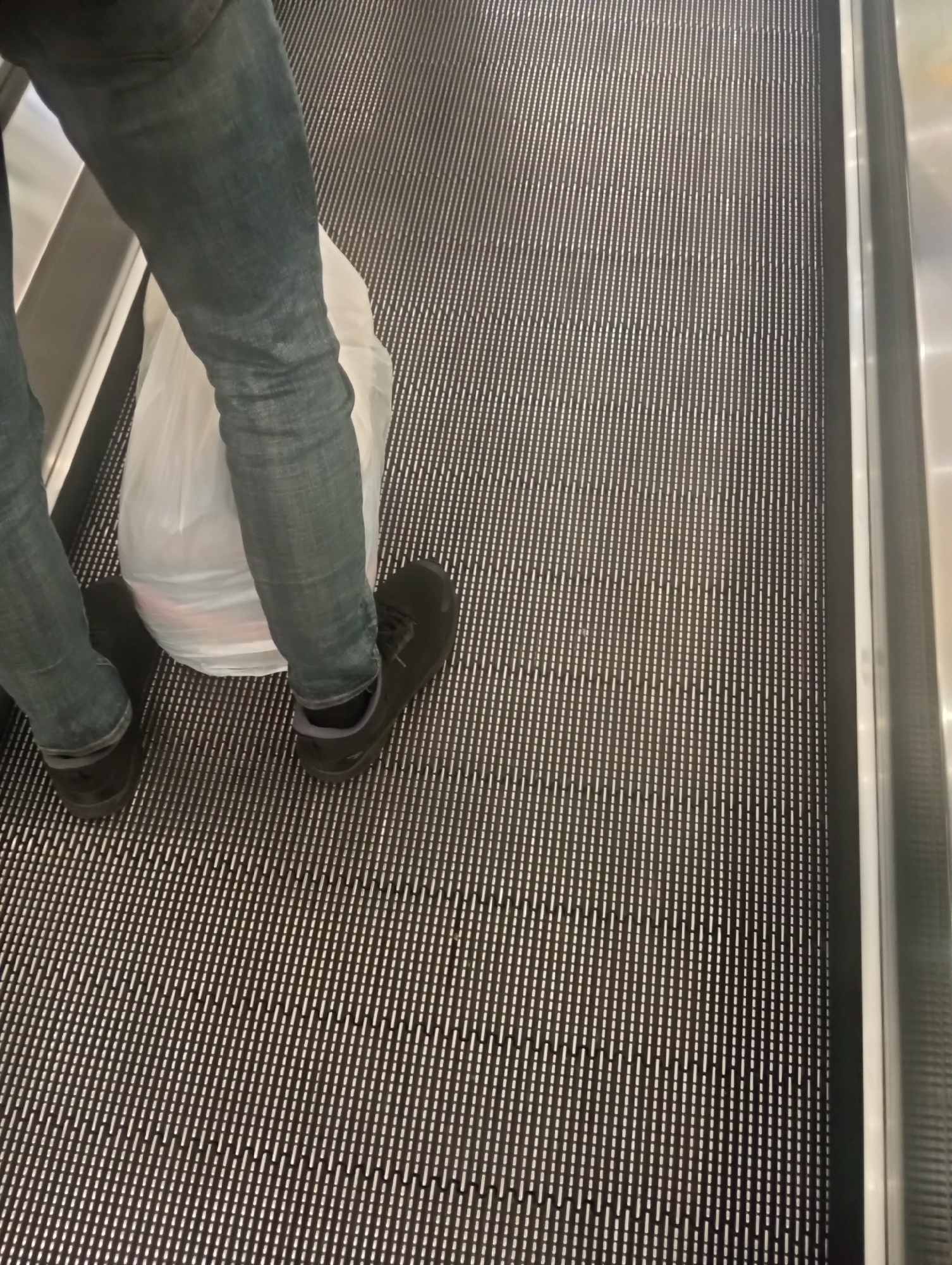 The image shows a person standing on an escalator in Surabaya, Indonesia. The person is wearing blue jeans and black shoes, and is holding a white plastic bag. The escalator steps are made of metal with a ribbed pattern. The background is blurred, but appears to be an indoor setting.