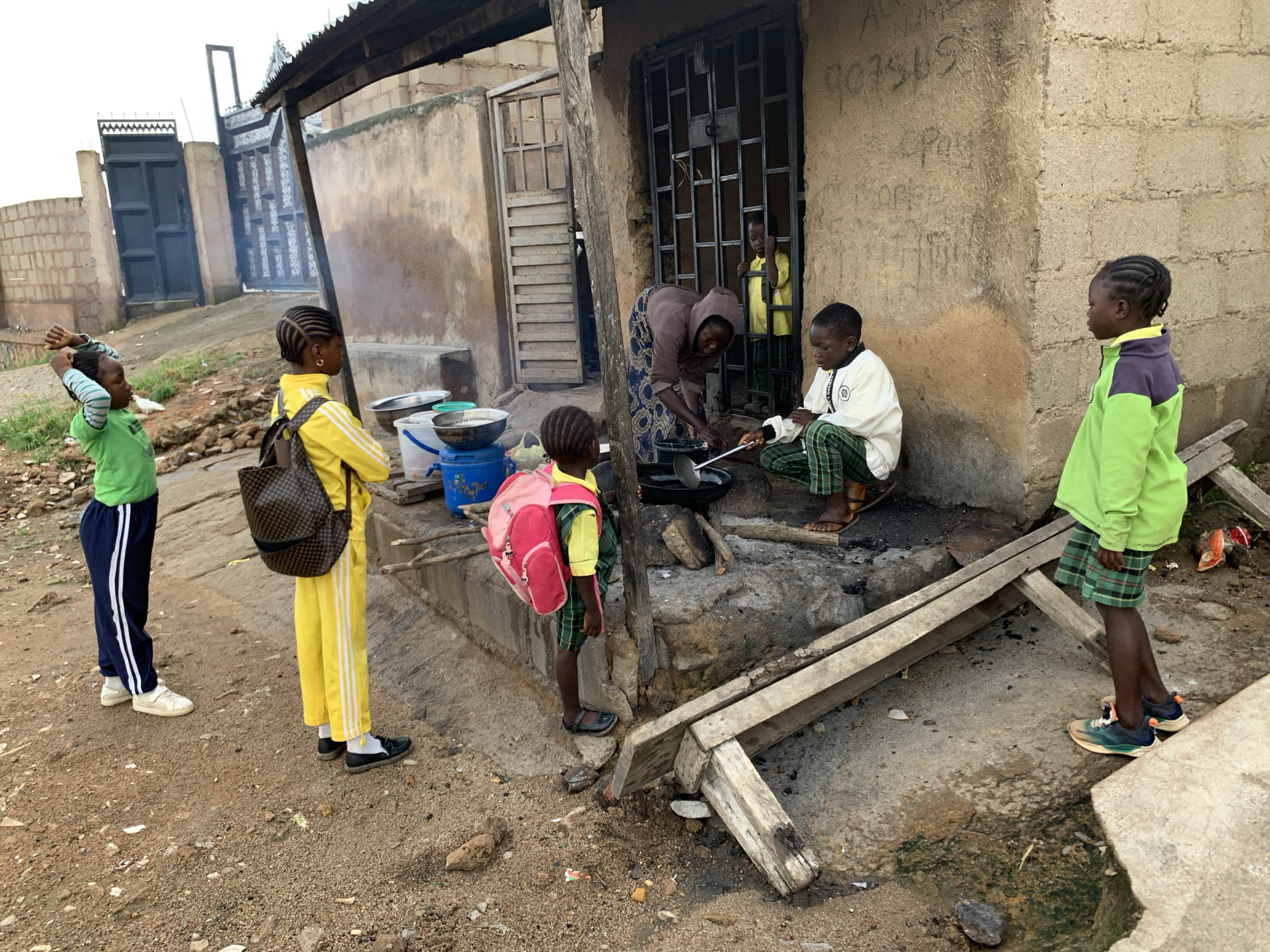 A group of children are gathered outside a building with a corrugated metal roof and a rough, concrete-block wall. One child, wearing a green long-sleeved shirt and blue pants with white stripes, stands to the left, arms raised. Next to them, a child in a yellow tracksuit and a patterned backpack stands facing away from the camera. A woman in a patterned dress and headscarf is kneeling near a cooking fire, stirring something in a pan with a ladle. To her right, a child in a white shirt and green-and-black checkered pants sits watching. Another child, wearing a bright pink backpack and a yellow and green outfit, stands facing the woman. On the far right, a child in a light green long-sleeved shirt and green plaid shorts stands looking towards the building. The ground is dirt and gravel, with some puddles of water. There are buckets and bowls near the cooking area. The building has a metal-barred door. Graffiti is visible on the wall of the building, including numbers that appear to be "907569". The scene suggests a communal outdoor gathering, possibly for cooking or preparing food, in a rural or semi-urban setting in Jos, Nigeria. The lighting suggests it is daytime.