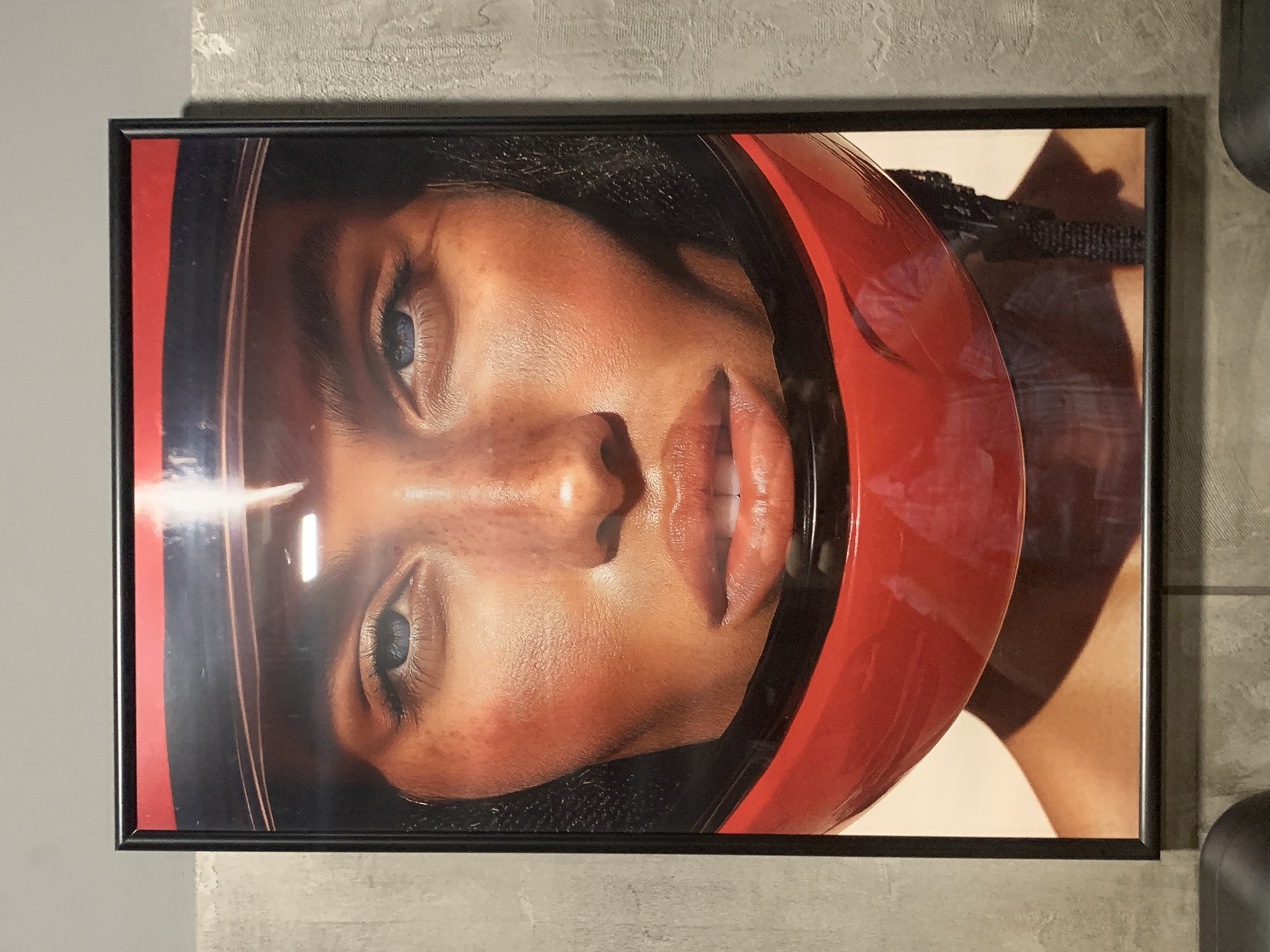 A framed portrait of a person wearing a red helmet and a black visor is displayed on a textured gray wall. The portrait is of a person's face, from just above the eyebrows to the chin, with the red helmet encircling the lower part of their face and head. The person has light blue eyes and light-colored skin with freckles. The lighting in the image is bright and appears to highlight the person's cheekbones and nose. The background of the portrait is a light peach color, and the framing is black. The wall behind the portrait has a rough, concrete-like texture. The overall impression is one of a close-up, artistic photograph.