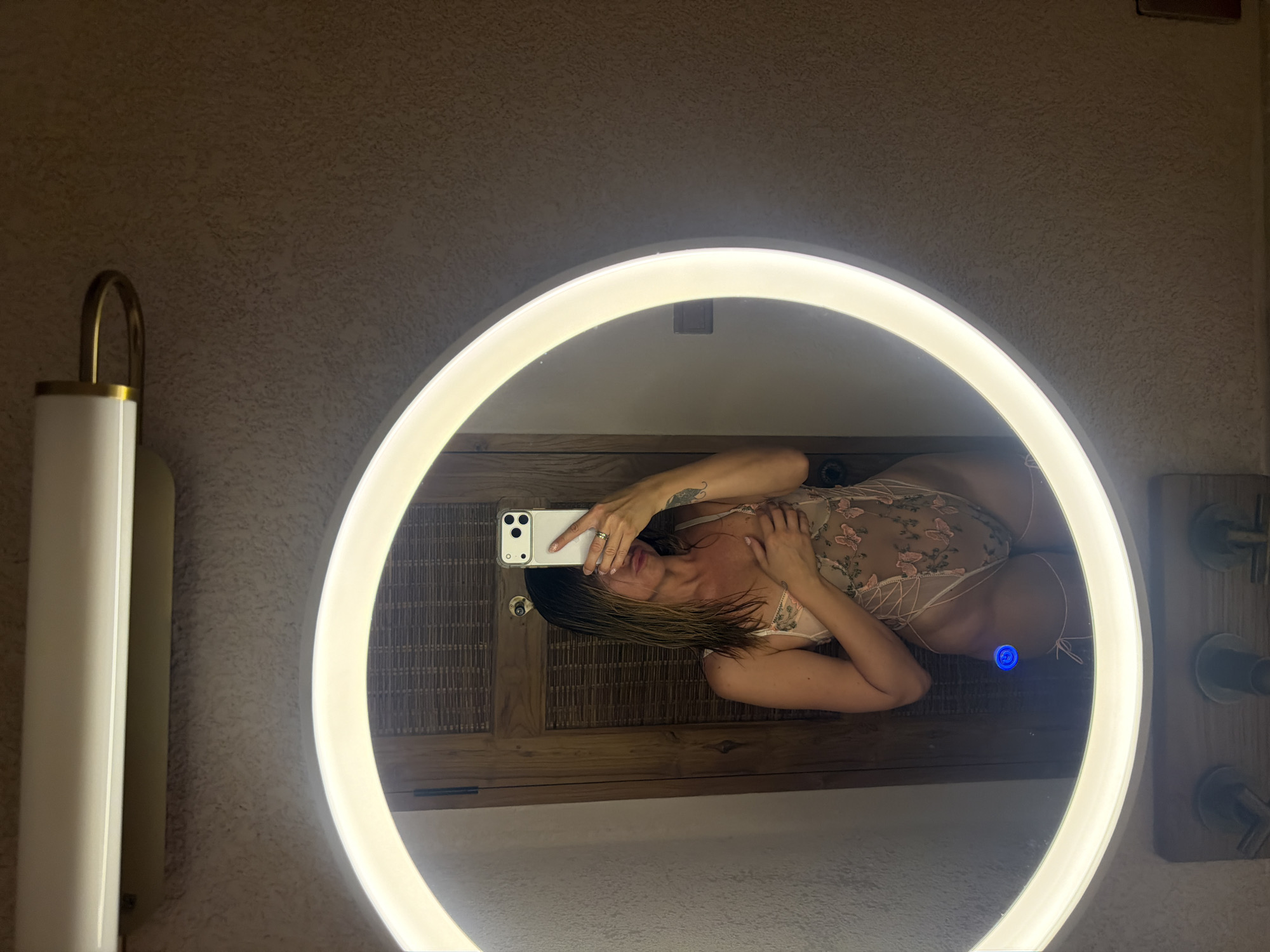A woman is taking a mirror selfie in what appears to be a bathroom. She is wearing a light-colored, sheer teddy with floral embroidery and criss-cross detailing. Her hand is placed over her chest, and she is holding a smartphone to take the picture. The mirror is circular with a bright LED light around its edge. Behind her, a wooden door with a woven texture is visible. The wall texture is rough and speckled. Below the mirror, what seems to be bathroom fixtures are partially visible. The scene is likely within a private residence or a hotel in Mengwi, Indonesia.