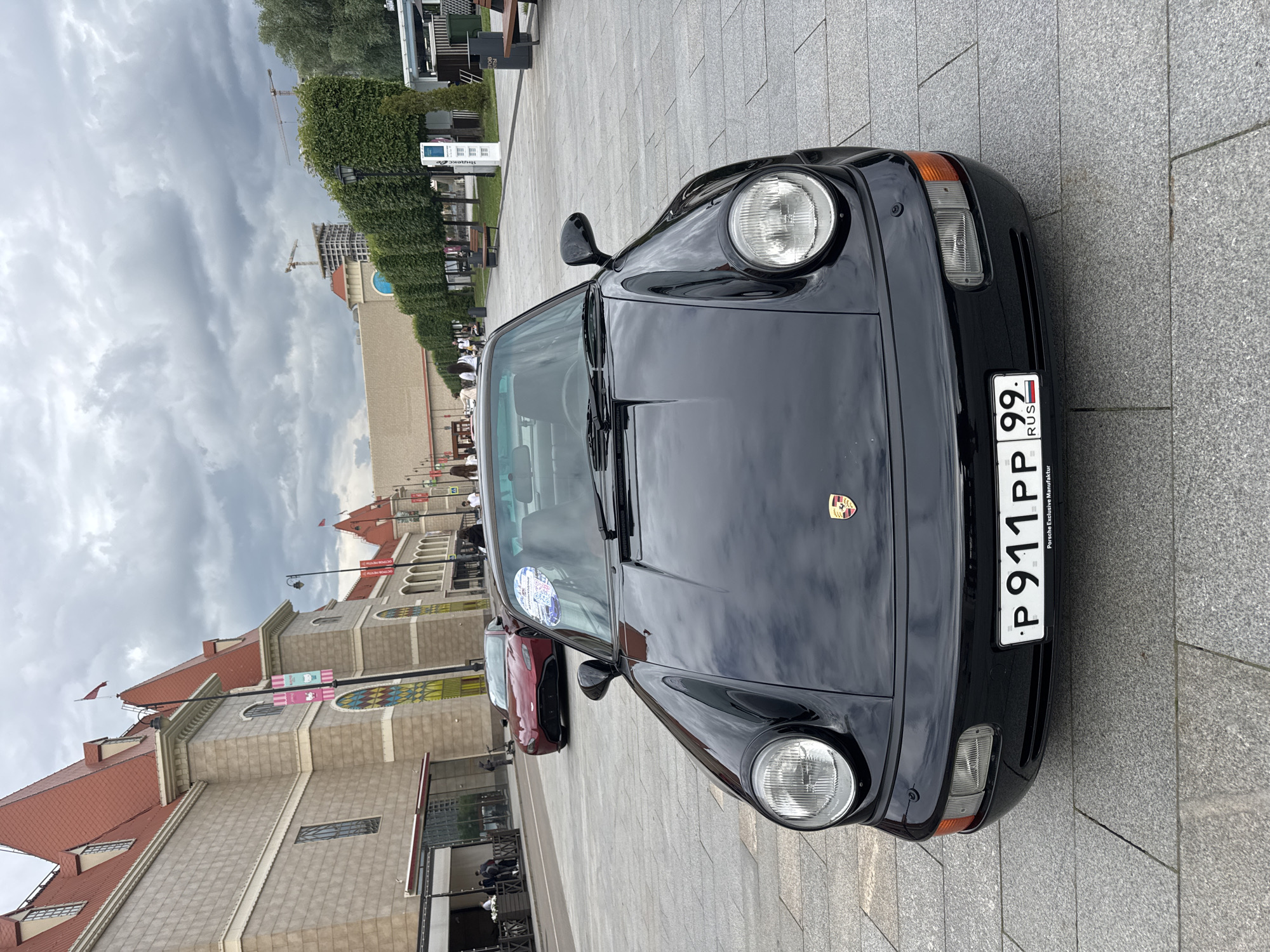 In Moscow, Russia, a black Porsche 911 is parked on a paved plaza in front of a building with a red tiled roof and ornate architecture. The car faces the camera, its headlights illuminated. A red sports car is visible in the background. The plaza is lined with trees and benches, and several people are walking in the distance. The sky is cloudy. The license plate on the Porsche reads "P 911 PP 99" and "RUS". The building appears to be a commercial or entertainment venue, possibly a shopping mall or exhibition center, with large windows and colorful banners. Construction cranes are visible in the far background, suggesting ongoing development in the area.