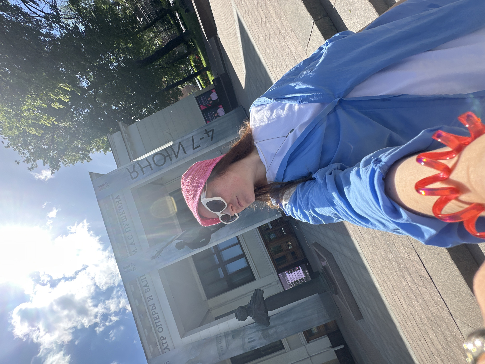 A person is captured in a sunlit selfie, positioned in the foreground with an outstretched arm. The individual wears a pink knitted hat, white sunglasses, a light blue long-sleeved shirt over a white top, and a distinctive red coiled bracelet on their right wrist. Their light brown hair is visible underneath the hat.
The background features a grand, light-colored building, likely a cultural institution, with a large, dark statue or bust in front of it. The building's facade displays Russian text and a graphic of a string instrument, possibly a cello or double bass. The sky is bright blue with scattered white clouds, and direct sunlight casts strong shadows, indicating daytime. Lush green trees are visible in the upper part of the frame.
Visible Russian text on the building includes "АТР ОПЕРЫ И БАЛЕТА" (Theatre of Opera and Ballet), "М. А. С. ПУШКИНА" (A. S. Pushkin, suggesting the institution is named after the poet), and "4-7 ИЮН" (June 4-7), possibly indicating event dates. Part of the word "ВНОМ" is also discernible. Given the context of Nizhny Novgorod, Russia, this is likely the A.S. Pushkin Nizhny Novgorod State Academic Opera and Ballet Theatre. The scene depicts a casual outdoor moment on a bright, clear day.