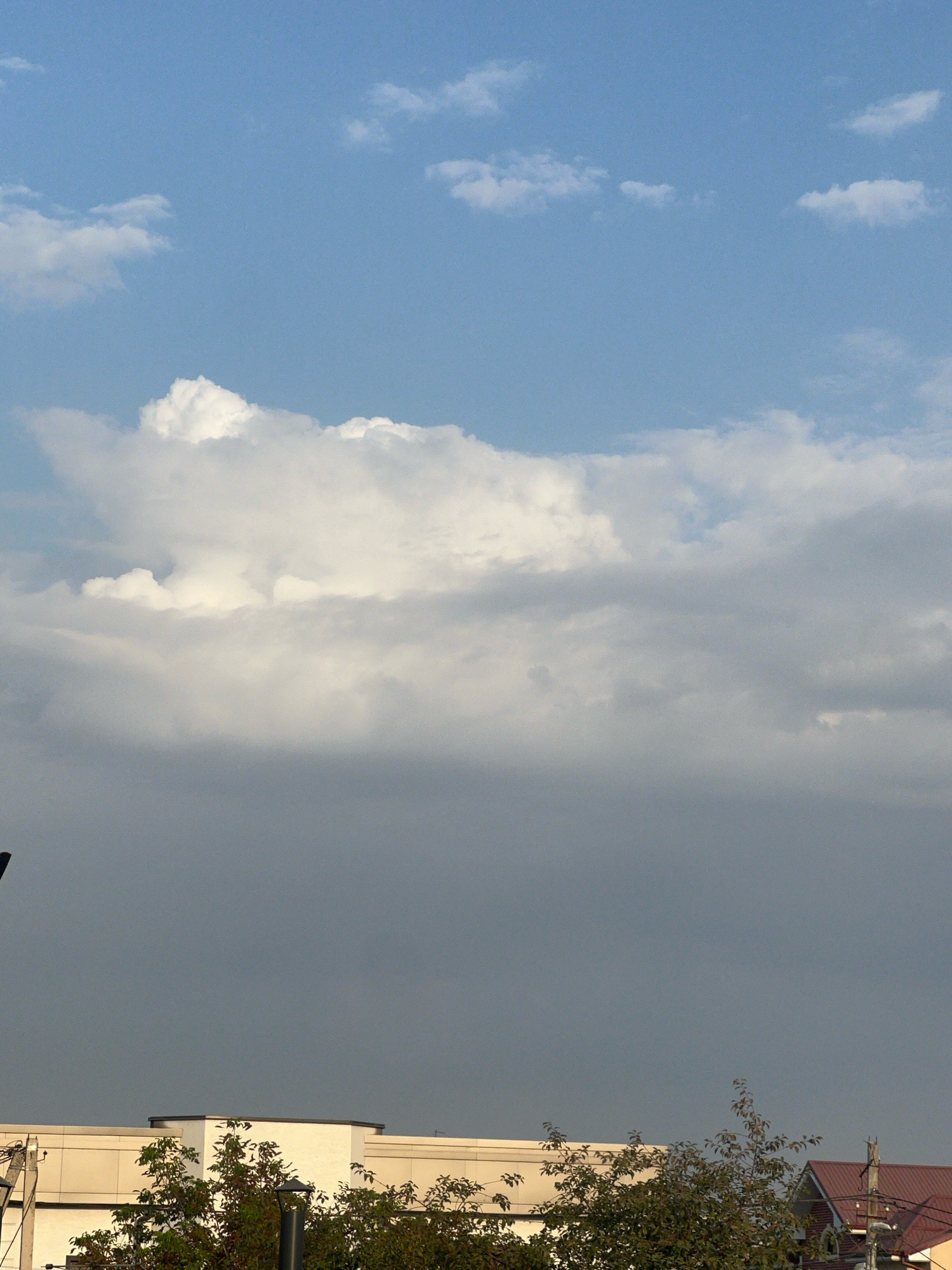 The image shows a view of the sky above buildings in a city. The sky is a mix of blue and gray, with various cloud formations. In the upper portion of the image, there are bright white cumulus clouds against a clear blue sky. Below these, a band of darker, gray clouds spans across the middle of the frame. The bottom of the image reveals the tops of buildings and trees. The buildings appear to be modern, with light-colored facades and dark trim. Trees with green foliage are visible in front of and around the buildings. The overall impression is of an urban landscape under a dynamic sky.